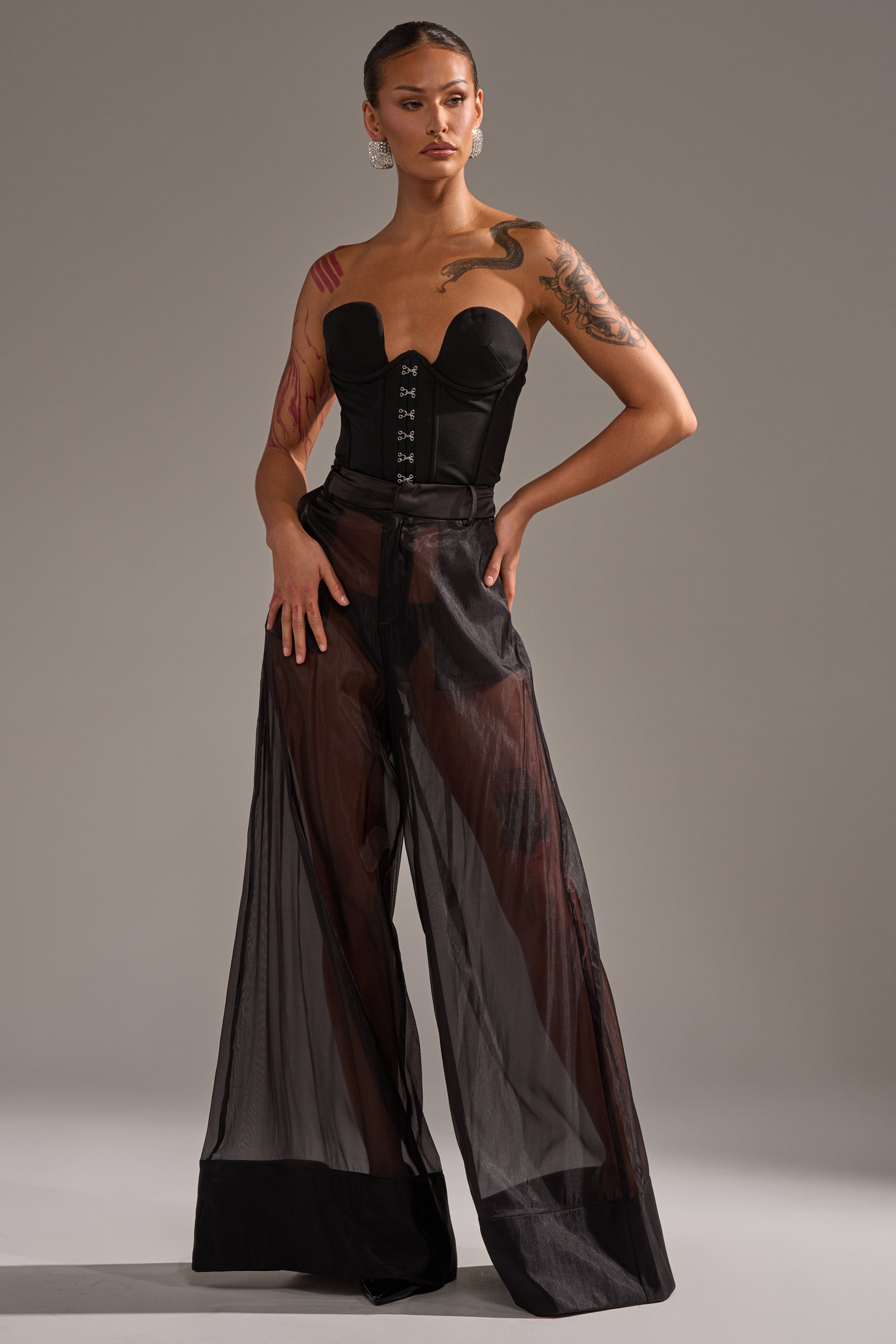 A tattooed person wears a black strapless corset top and the LOUD AND LUX TROUSER—sheer, wide-leg black pants—posing confidently against a plain gray background. 5