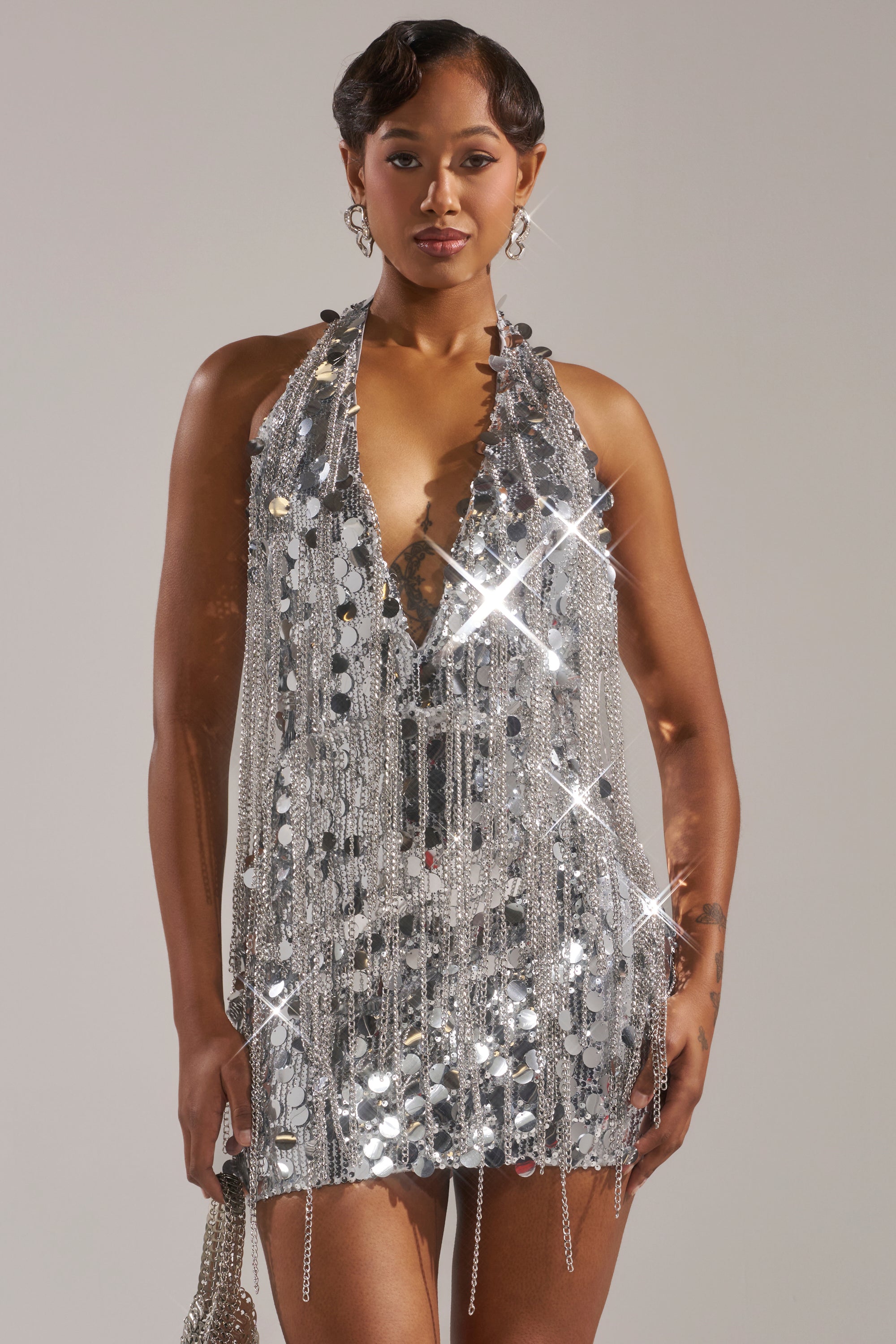 A woman faces forward in the LATE NIGHT AT THE DISCO SEQUIN MINI DRESS—a sleeveless silver sequin mini with a plunging neckline—dazzling onlookers as she holds a matching silver bag.