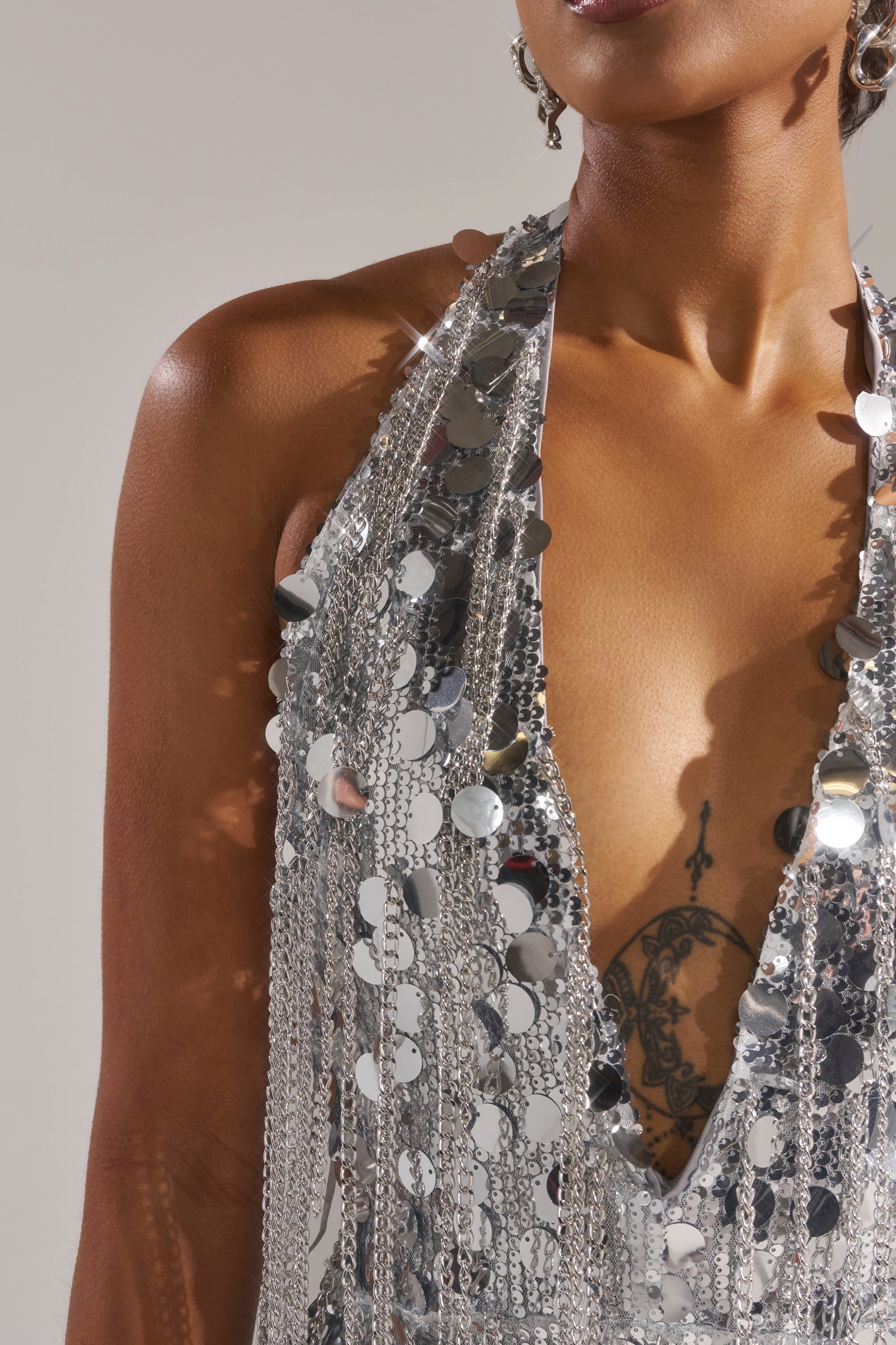 A person wears the LATE NIGHT AT THE DISCO SEQUIN MINI DRESS, a shiny silver halter mini with a deep V neckline that reveals a butterfly chest tattoo, set against a plain, neutral background.