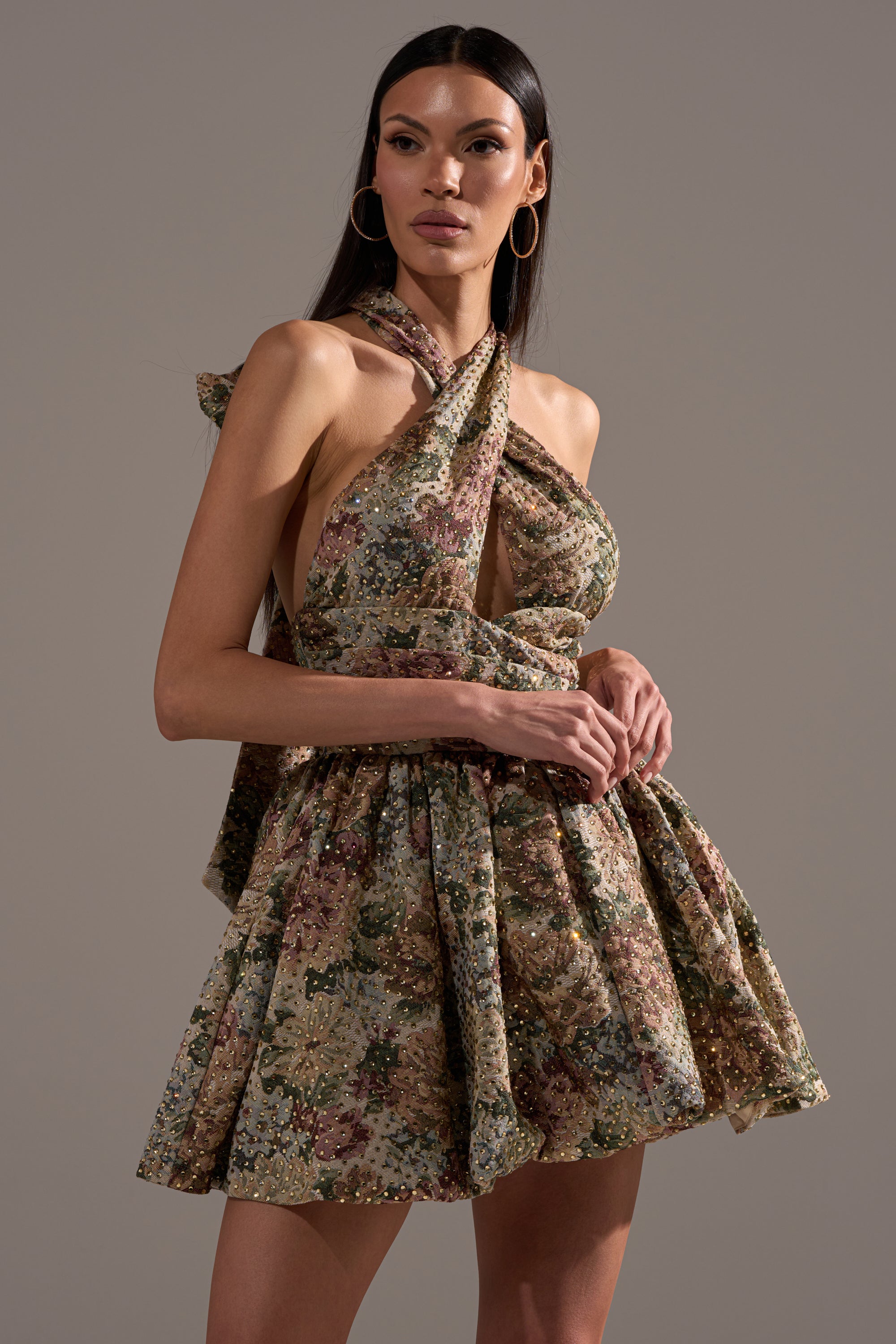 A woman with long straight hair and hoop earrings stands confidently against a gray background, wearing the GUESS WHO FLORAL MINI DRESS—a sleeveless halter-neck mini dress featuring a floral print and full bubble skirt.
