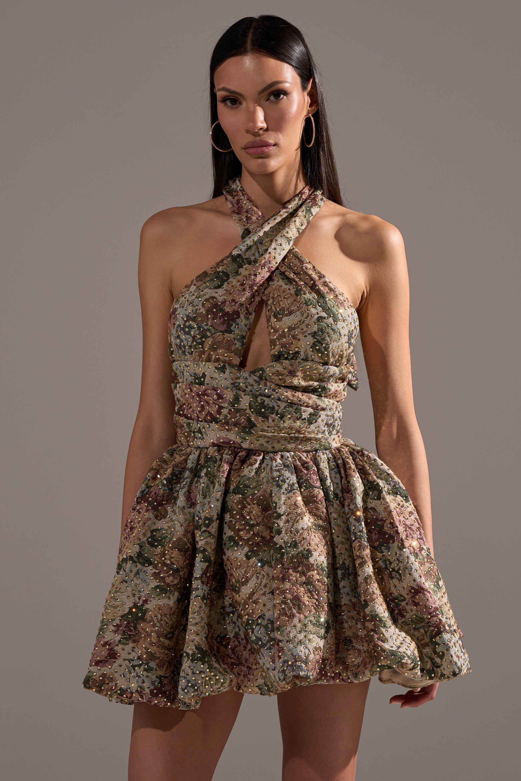 Woman wearing the GUESS WHO FLORAL MINI DRESS, featuring a crisscross halter neckline and chest cutout, stands against a neutral background—a true statement party piece.
