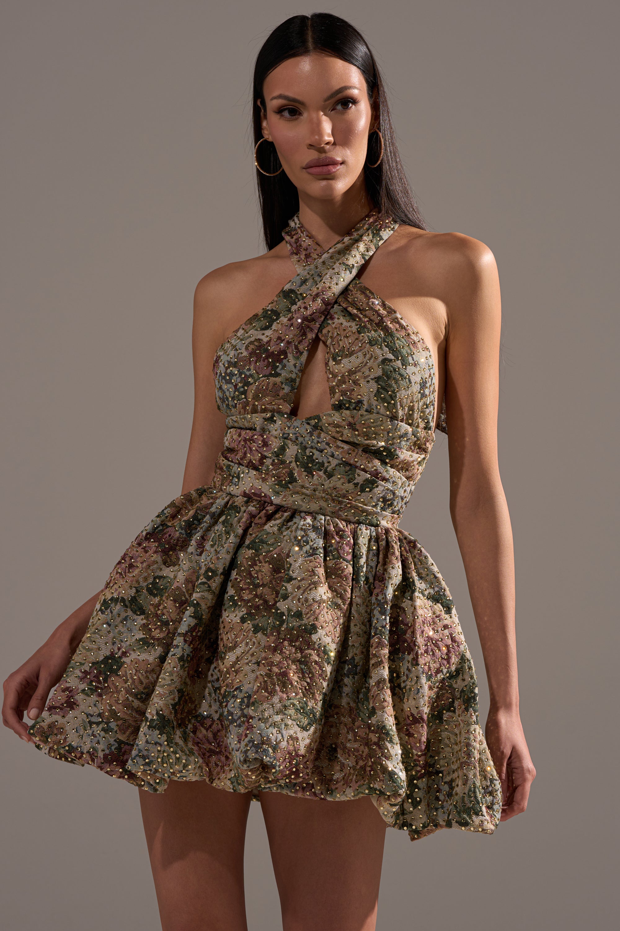 A woman poses against a gray background in the GUESS WHO FLORAL MINI DRESS, featuring a halter neck, crisscross bodice, and flared skirt—an ideal statement party look.