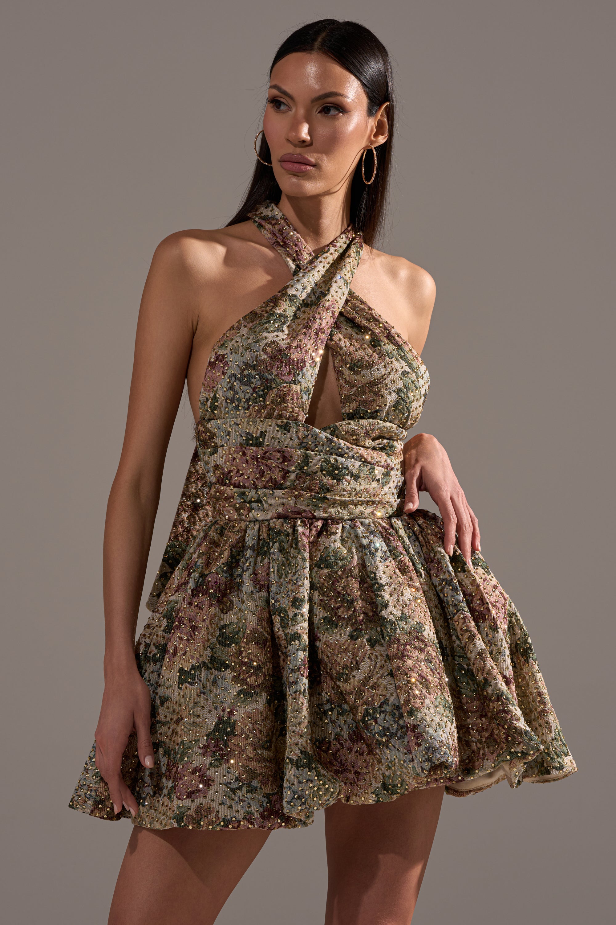 Wearing the GUESS WHO FLORAL MINI DRESS—featuring a halter neck, full skirt, and metallic accents—a woman poses against a plain background with one hand on her hip, making it perfect for special occasions.