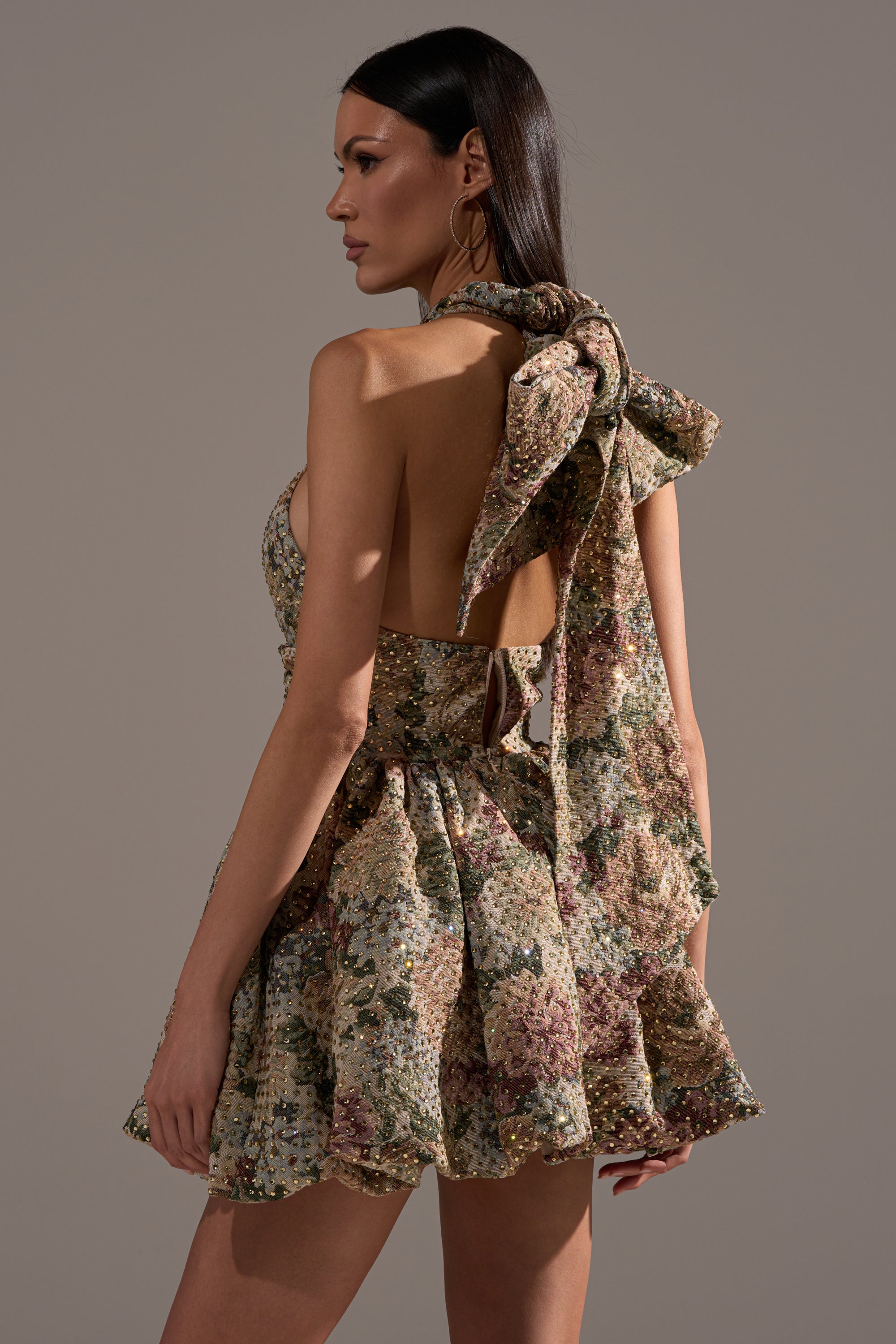 A woman models the GUESS WHO FLORAL MINI DRESS featuring a floral jacquard design, a statement bow on one shoulder, and an open back—ideal for any special occasion. She stands against a neutral background with straight brown hair.