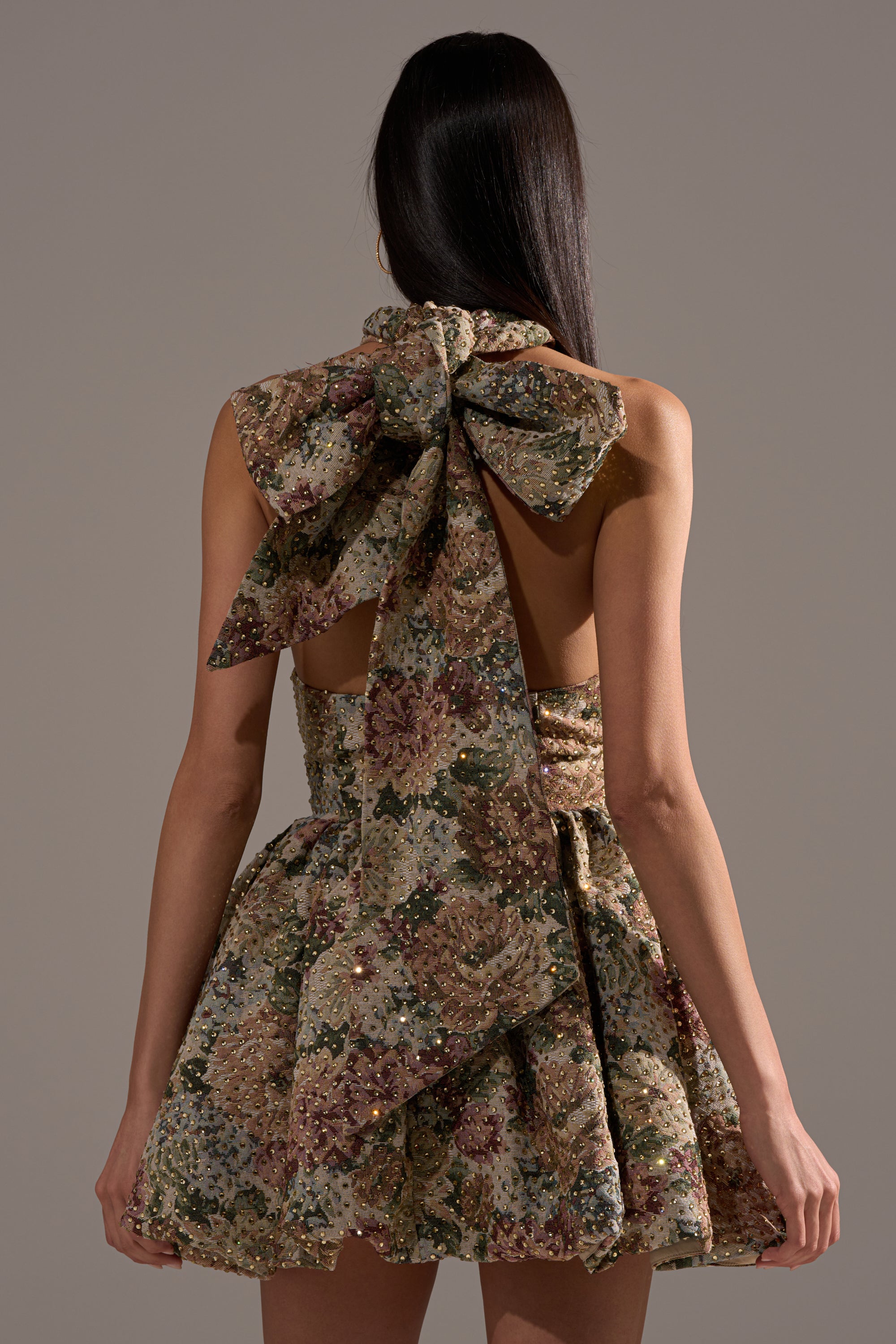 A woman with long, straight dark hair is seen from behind wearing the GUESS WHO FLORAL MINI DRESS, featuring a sleeveless cut, dramatic bow, open back, and muted earth tone florals—a statement party look.