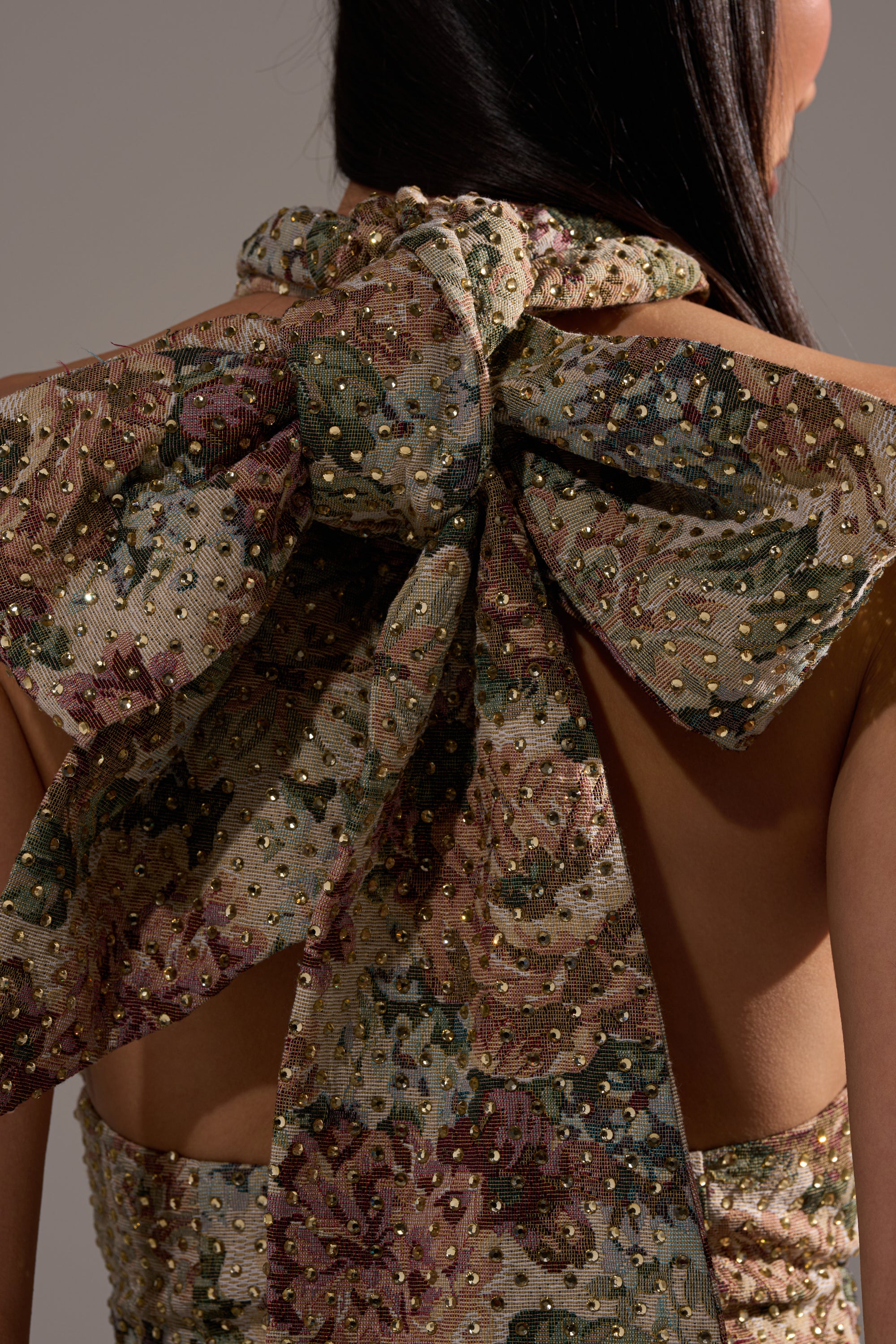 Close-up of a woman's back in the GUESS WHO FLORAL MINI DRESS, a jacquard party dress with an open back, large decorative bow, gold embellishments, and textured fabric in muted green, pink, and gold floral tones.