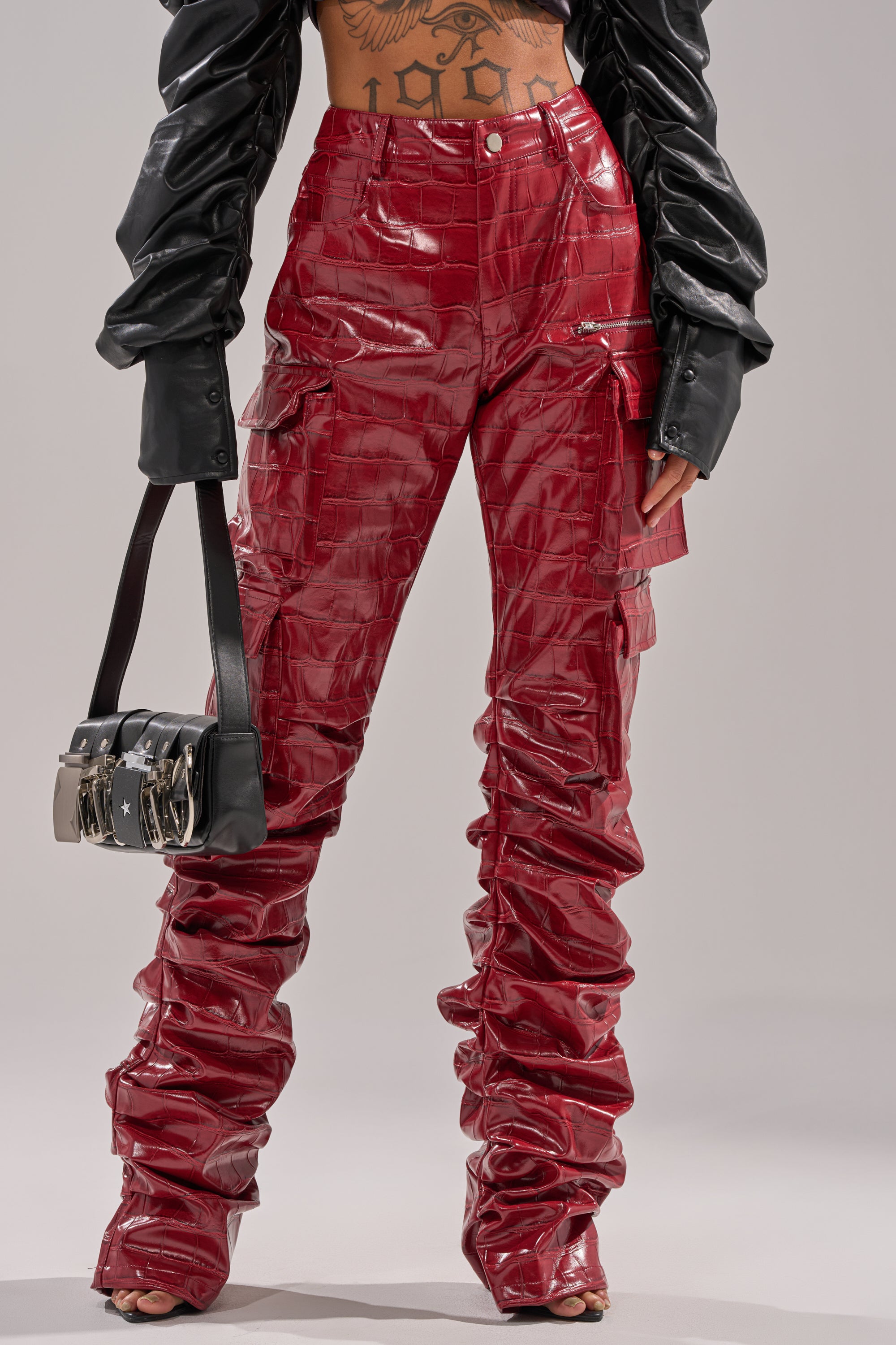 Wearing the STRAIGHT TO THE POINT STACKED FAUX LEATHER TROUSER—glossy, high-rise red pants with a ruched, stacked design and cargo pockets—paired with a black cropped jacket, handbag, and showing off a tattooed torso.