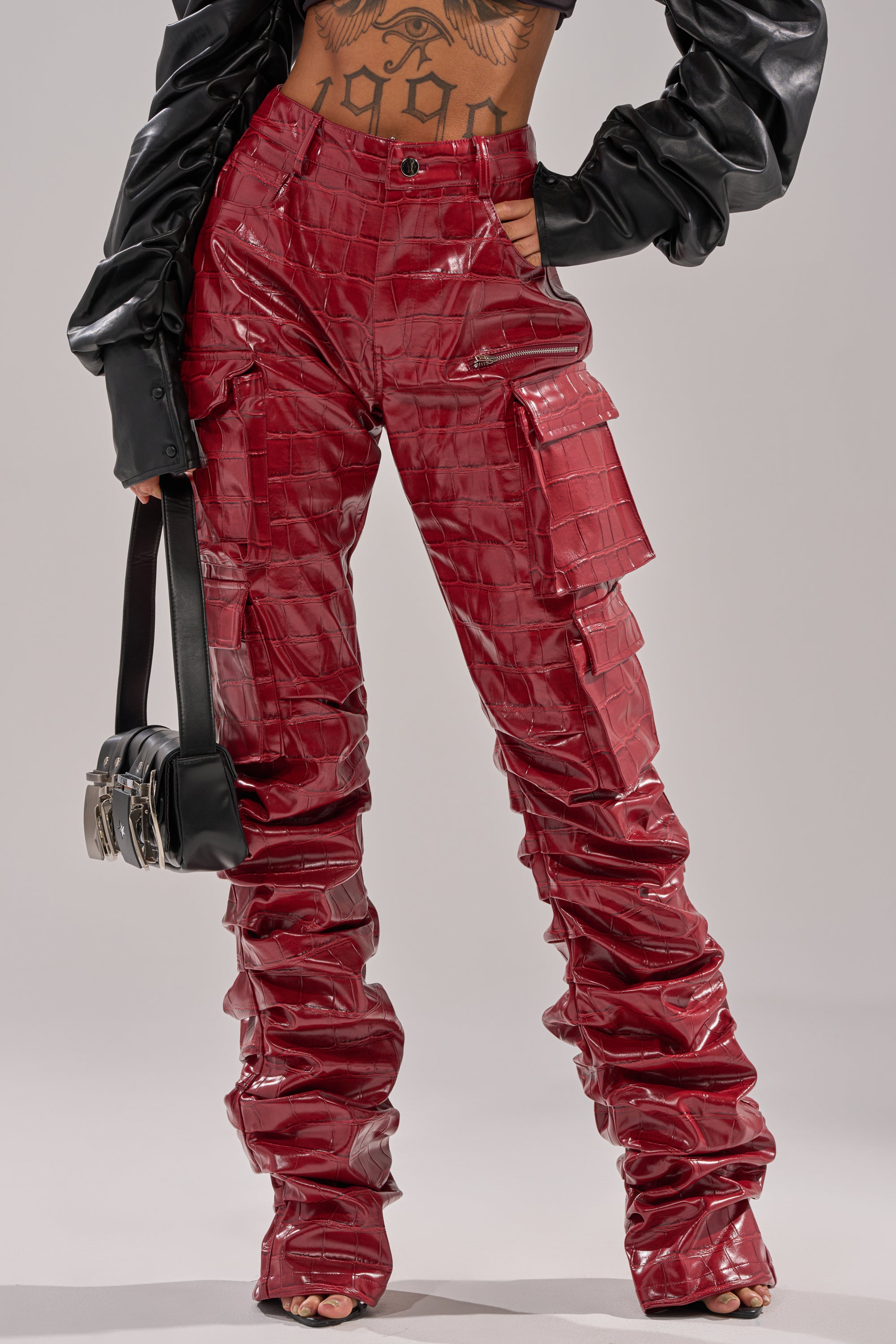 A person wearing the STRAIGHT TO THE POINT STACKED FAUX LEATHER TROUSER in shiny red, paired with a black long-sleeve puffed crop top and holding a small black handbag. The image shows only the body from the chest down.