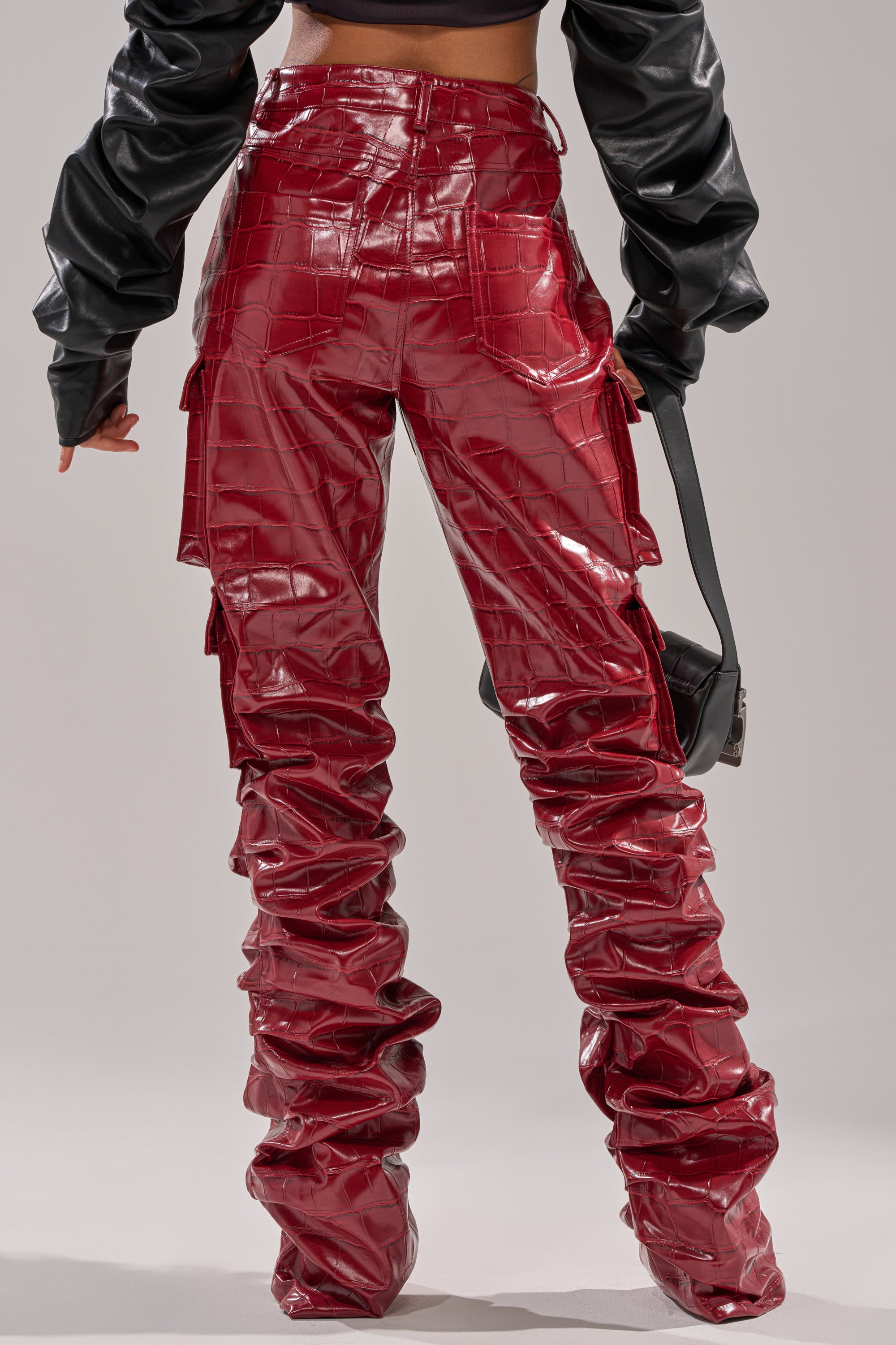 A person is seen from the back wearing the STRAIGHT TO THE POINT STACKED FAUX LEATHER TROUSER in glossy red crocodile-embossed faux leather with a ruched, high-rise stacked design, paired with a black cropped top and holding a black purse.