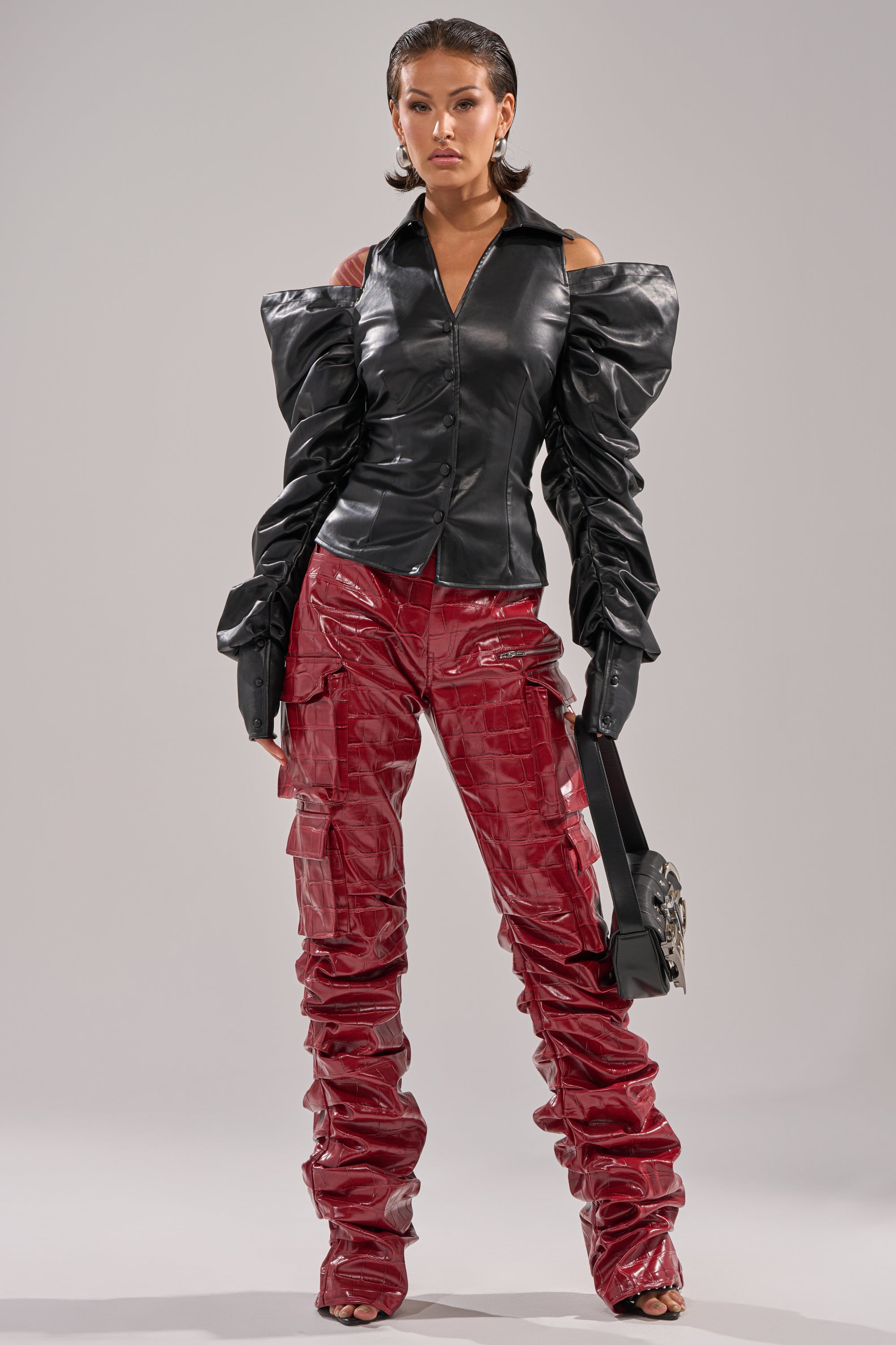 A woman poses confidently in a black off-shoulder faux leather top with voluminous sleeves and shiny, high-rise STRAIGHT TO THE POINT STACKED FAUX LEATHER TROUSER. She carries a black handbag and has sleek, wet-look hair.