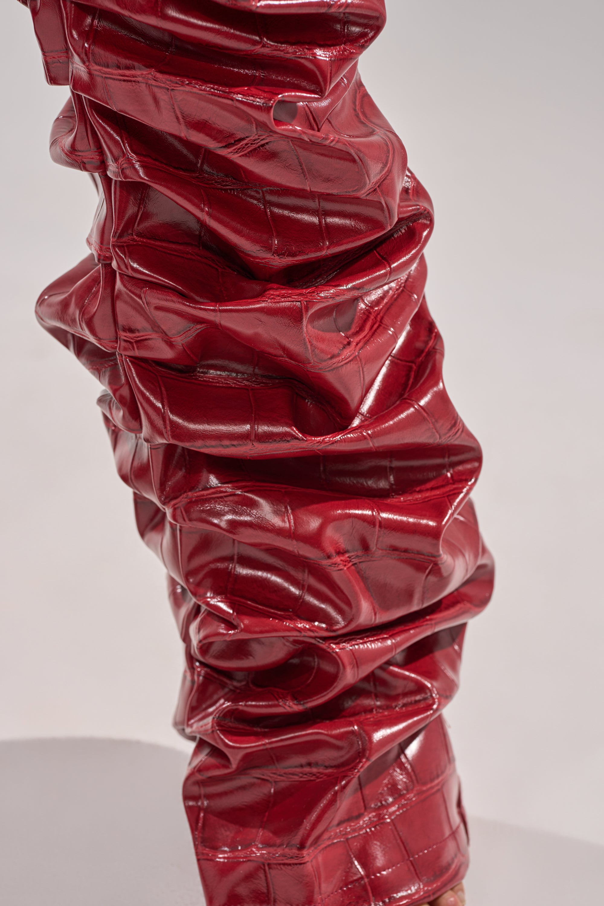 A close-up of the STRAIGHT TO THE POINT STACKED FAUX LEATHER TROUSER, featuring a shiny, textured red finish with a ruched, quilted pattern that mimics crocodile-embossed leather.