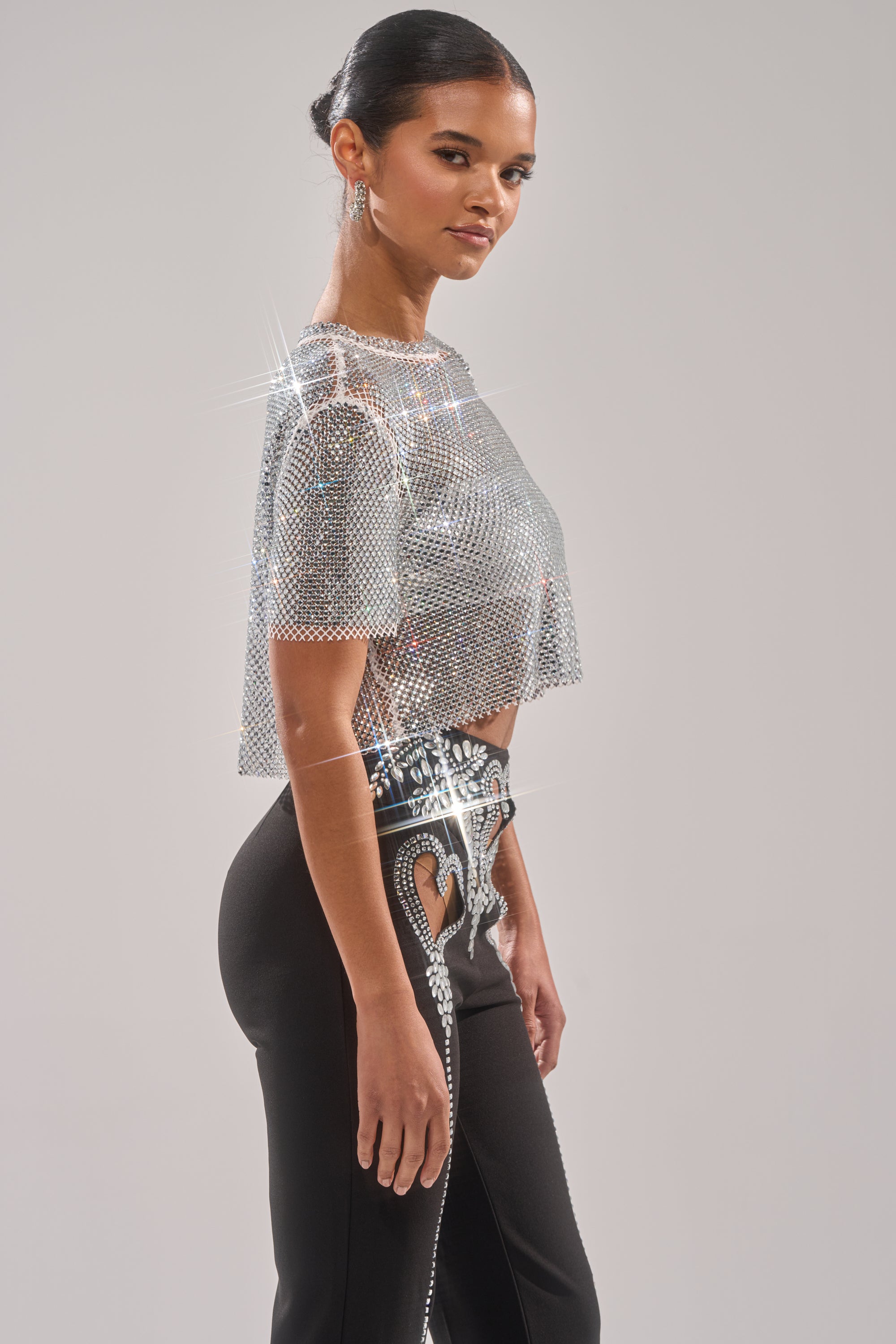 A woman exudes confidence in the JUST A FEELING RHINESTONE TEE, a sparkling silver fishnet top paired with high-waisted black pants featuring rhinestone details and cut-outs. Her hair is styled in a sleek bun.