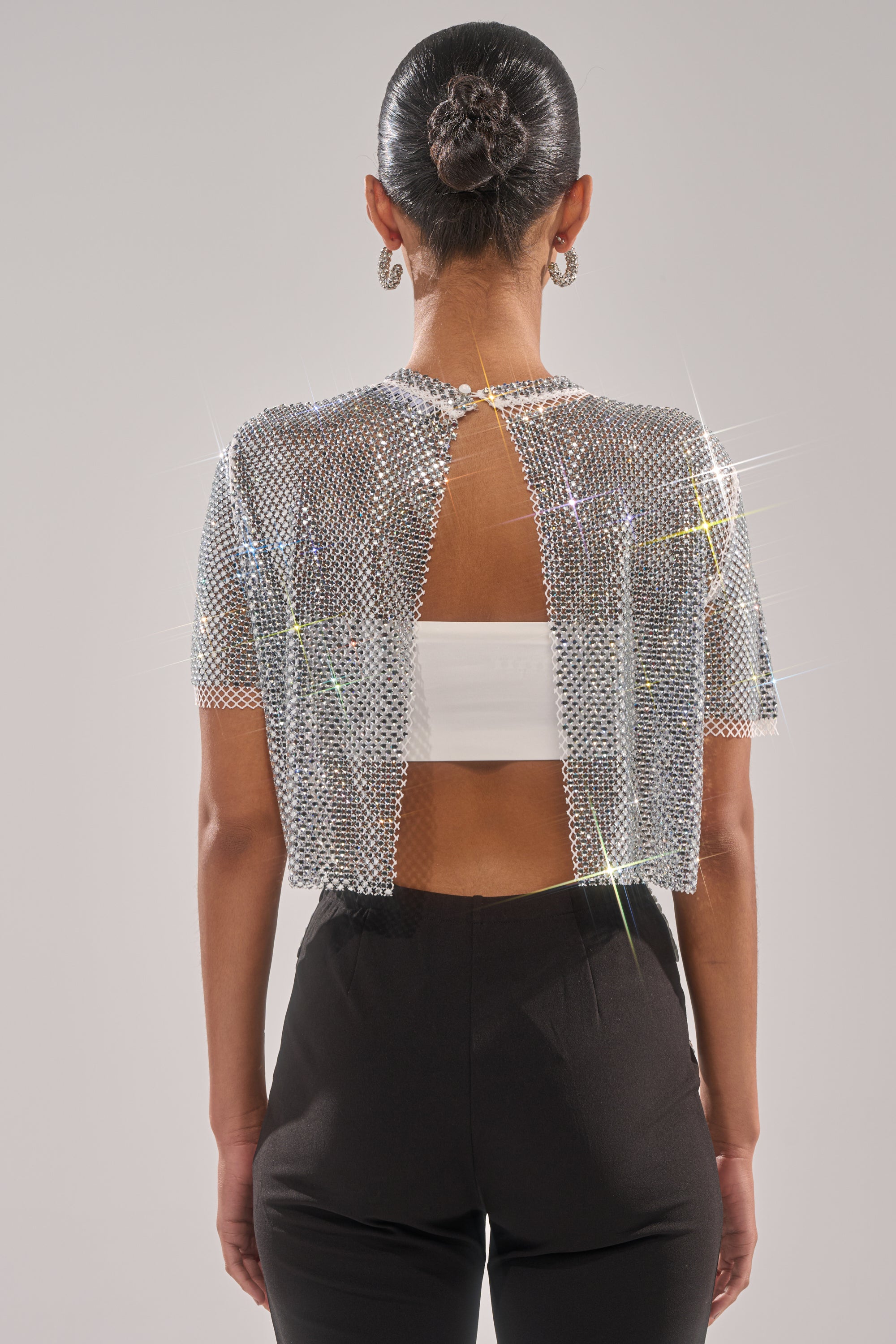 A woman faces away, wearing the JUST A FEELING RHINESTONE TEE with a cropped fit over a white bandeau and black high-waisted pants. She has a sleek bun and hoop earrings, standing against a plain, light background.