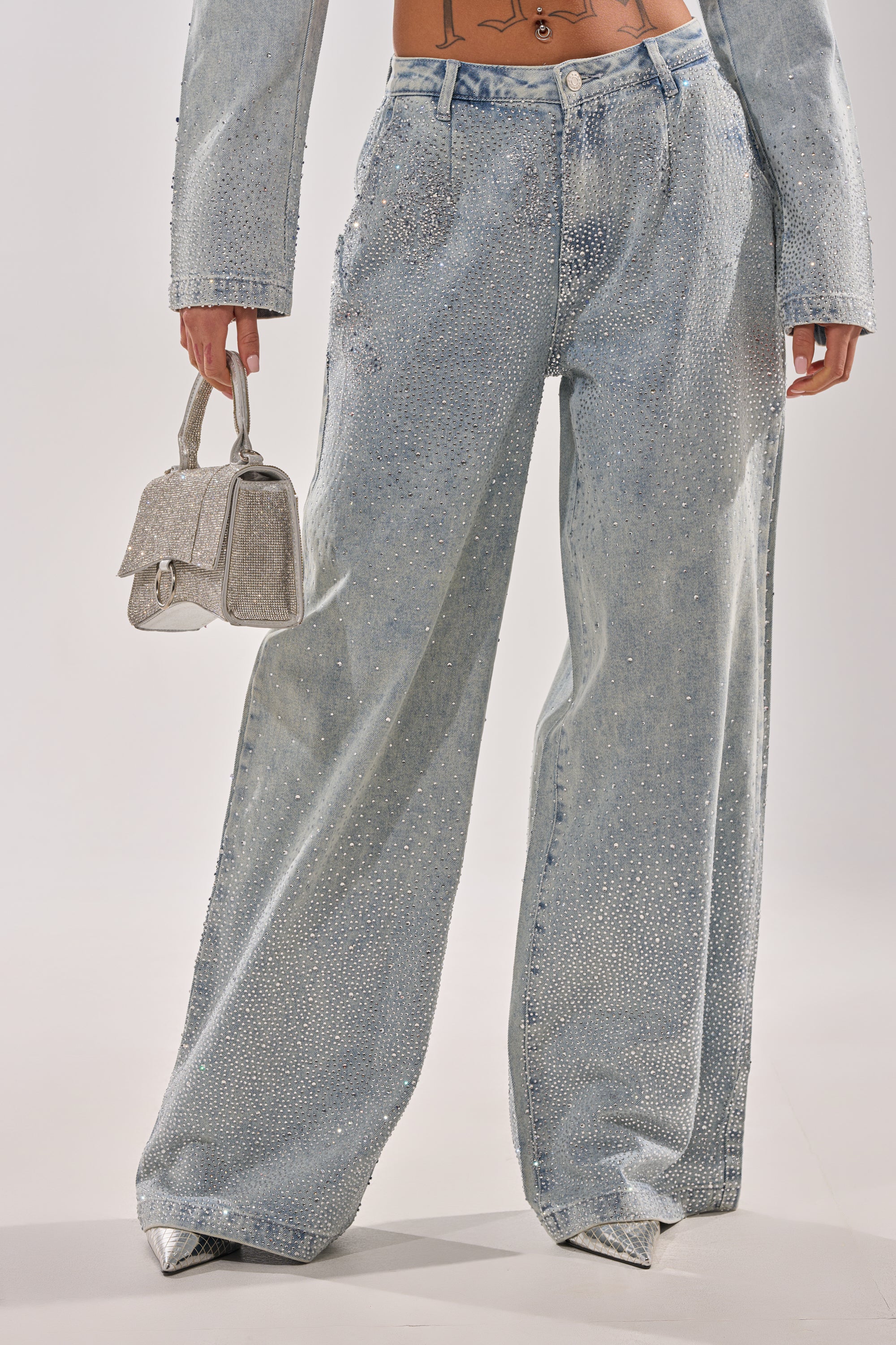 A person’s lower body is shown in the LIGHTS OUT RHINESTONE WIDE LEG DENIM and a matching jacket with rhinestones, styled with a sparkly silver handbag and silver pointed-toe heels. 1