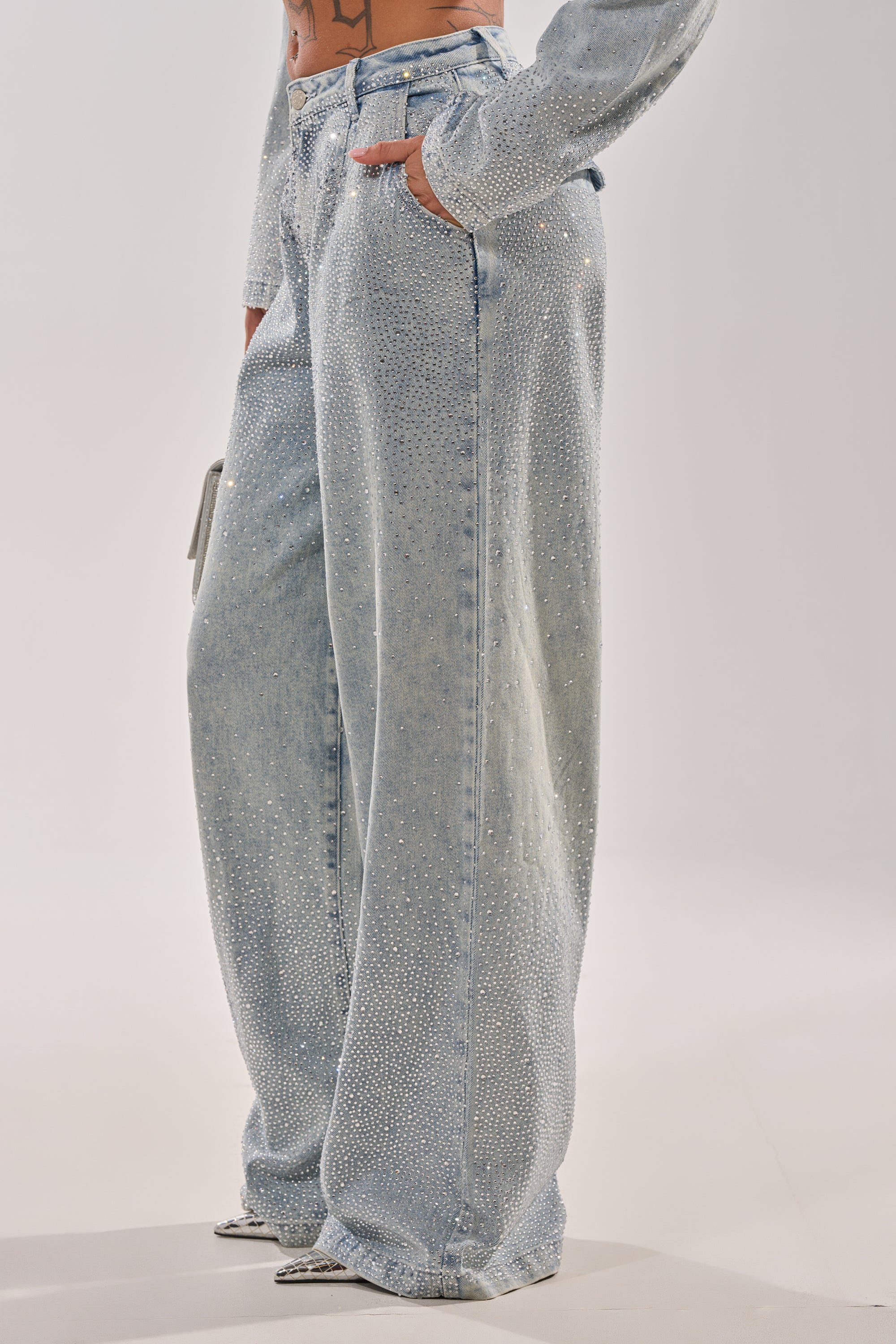 A person styles the LIGHTS OUT RHINESTONE WIDE LEG DENIM with a matching long-sleeve top and pointed-toe shoes, hands in pockets, for an on-trend look. 3