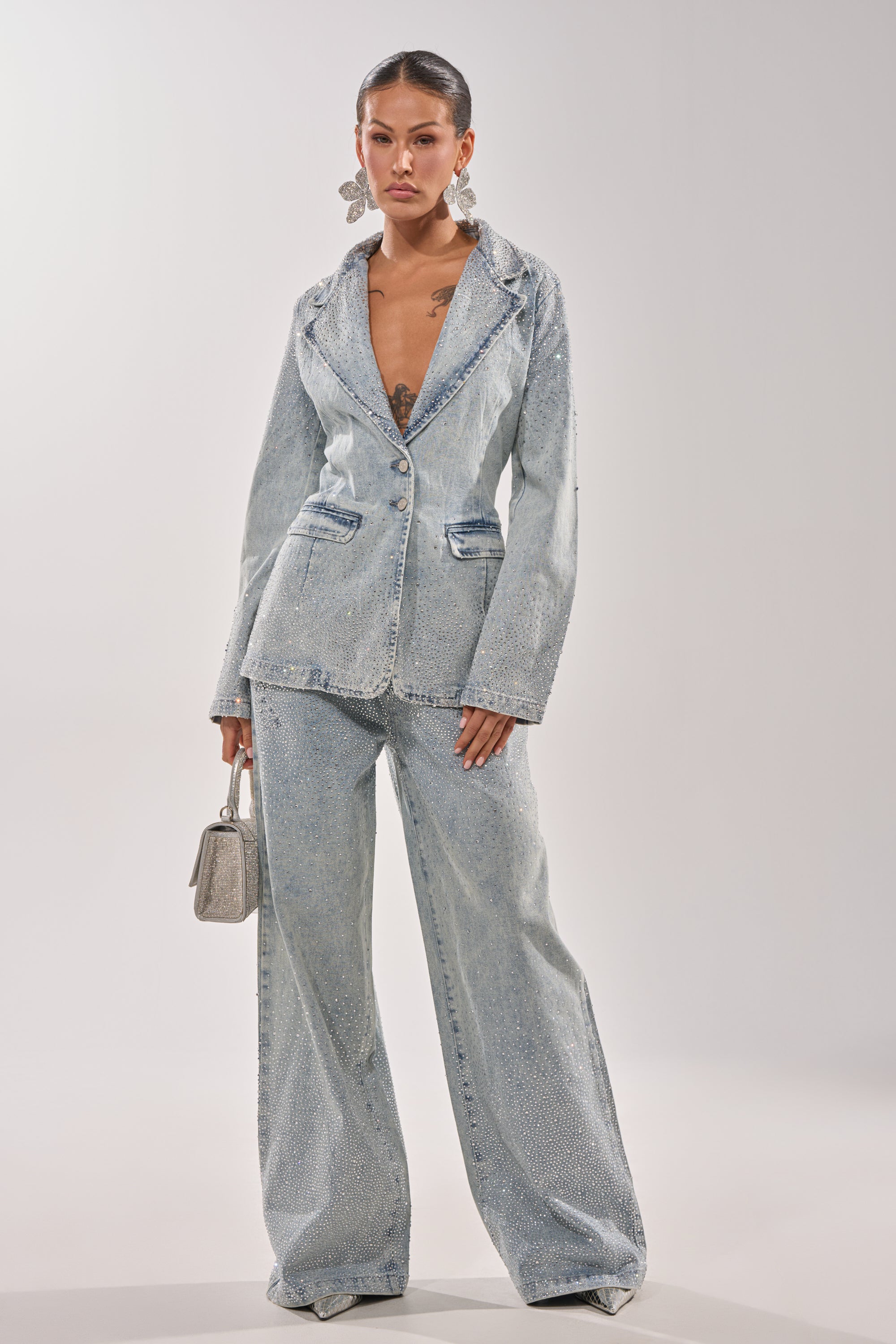 A woman stands confidently in LIGHTS OUT RHINESTONE WIDE LEG DENIM, paired with a light blue crystal-embellished blazer, statement earrings, and a small silver handbag. Her hair is pulled back against a plain light background. 5
