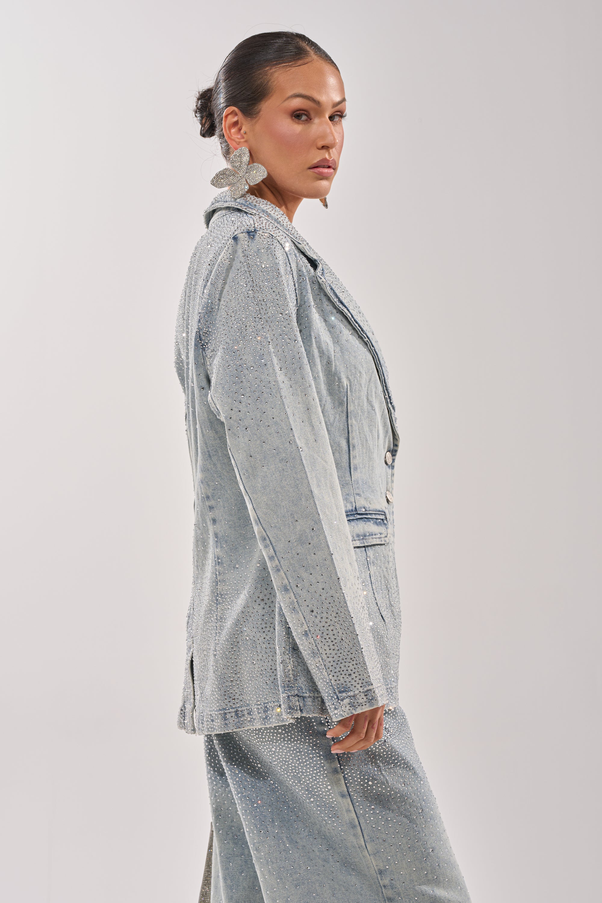 A woman with slicked-back hair models the LIGHTS OUT RHINESTONE DENIM BLAZER and matching pants with bold floral earrings, standing in profile against a white background—an elevated take on casual style. 3