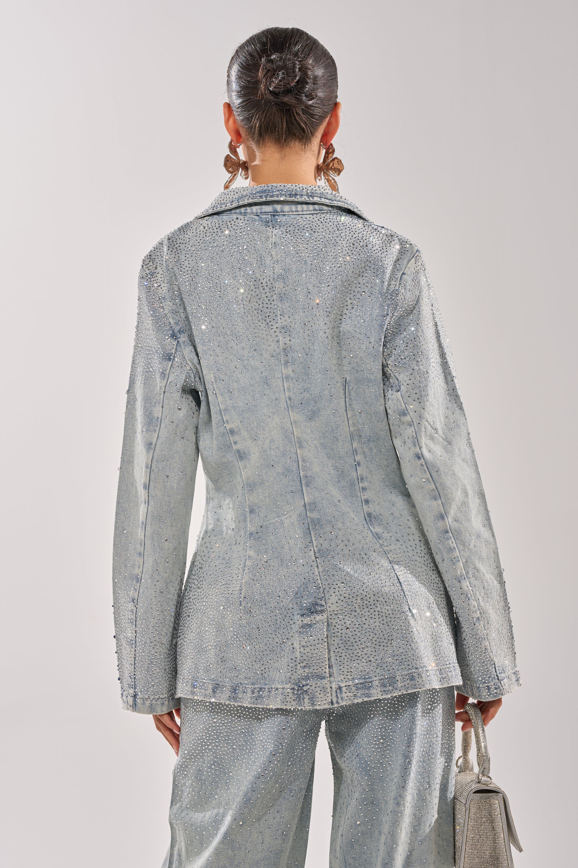 A person with dark hair in a bun is seen from the back wearing the LIGHTS OUT RHINESTONE DENIM BLAZER and matching pants, carrying a light handbag and accessorizing with large hoop earrings. 4