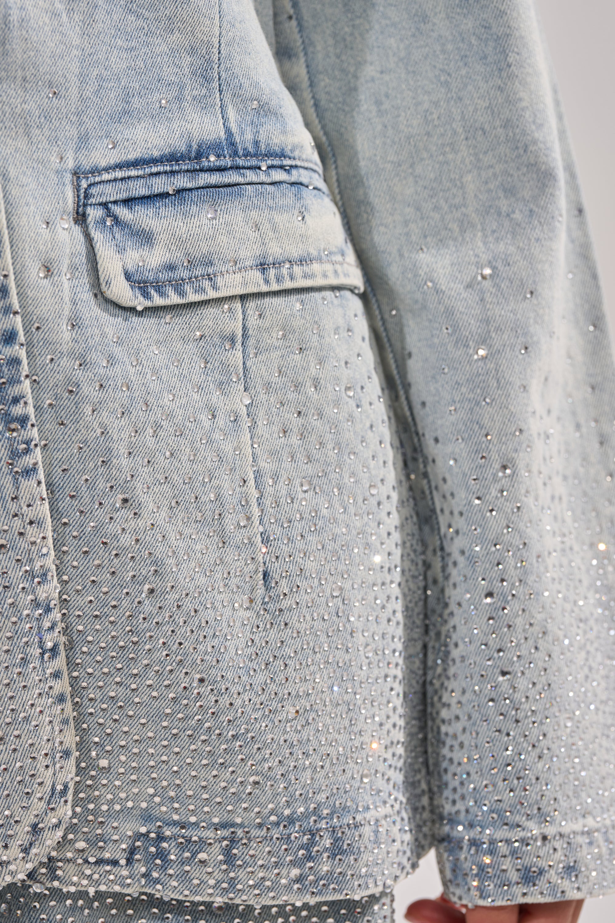 Close-up of the LIGHTS OUT RHINESTONE DENIM BLAZER in light blue, showcasing sparkling rhinestones, a pocket flap, and part of the sleeve. 7