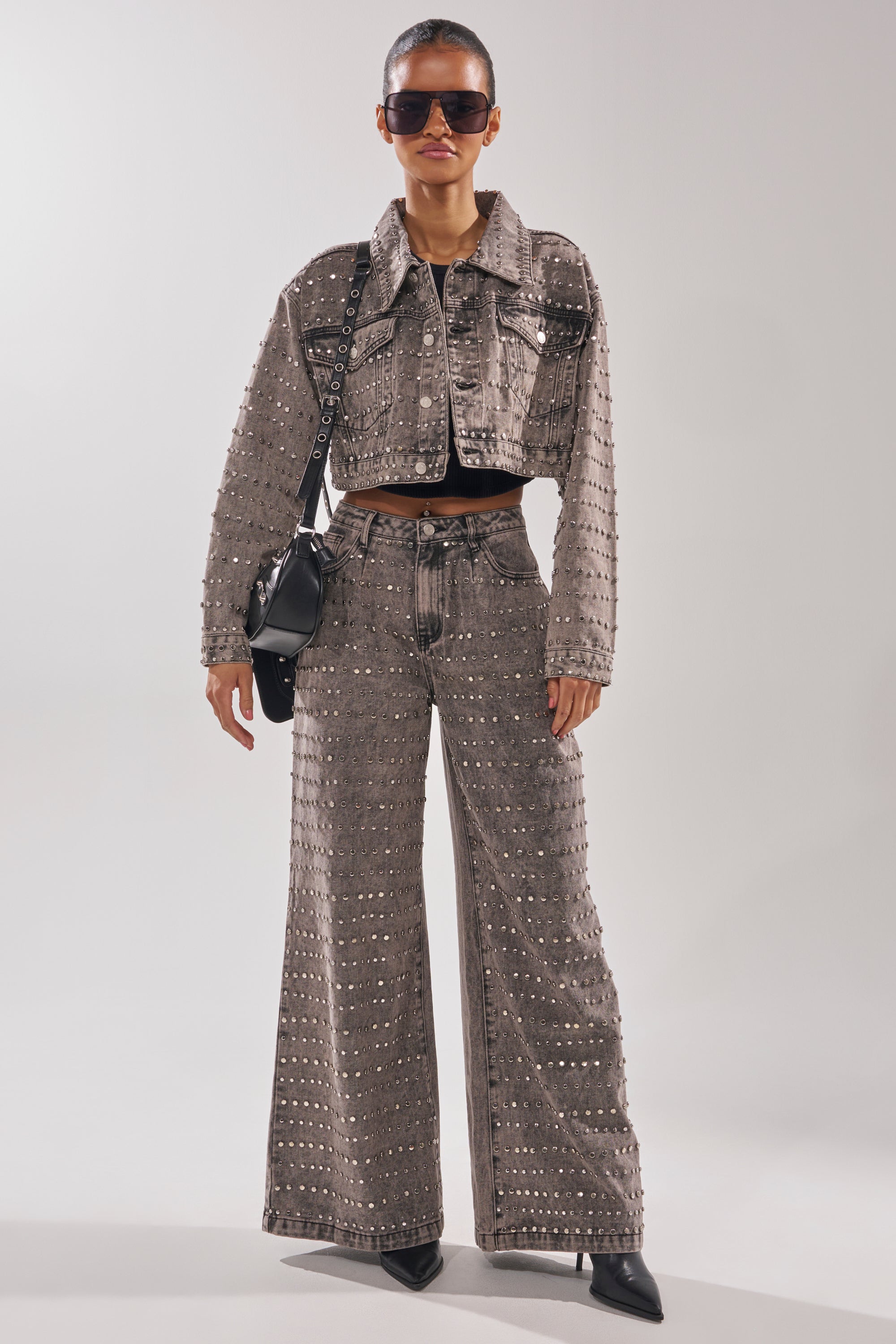 A woman stands confidently against a plain background in the RIZZO STUDDED WIDE LEG DENIM jacket and matching high waist jeans, styled with black pointed-toe shoes, sunglasses, and a black shoulder bag.