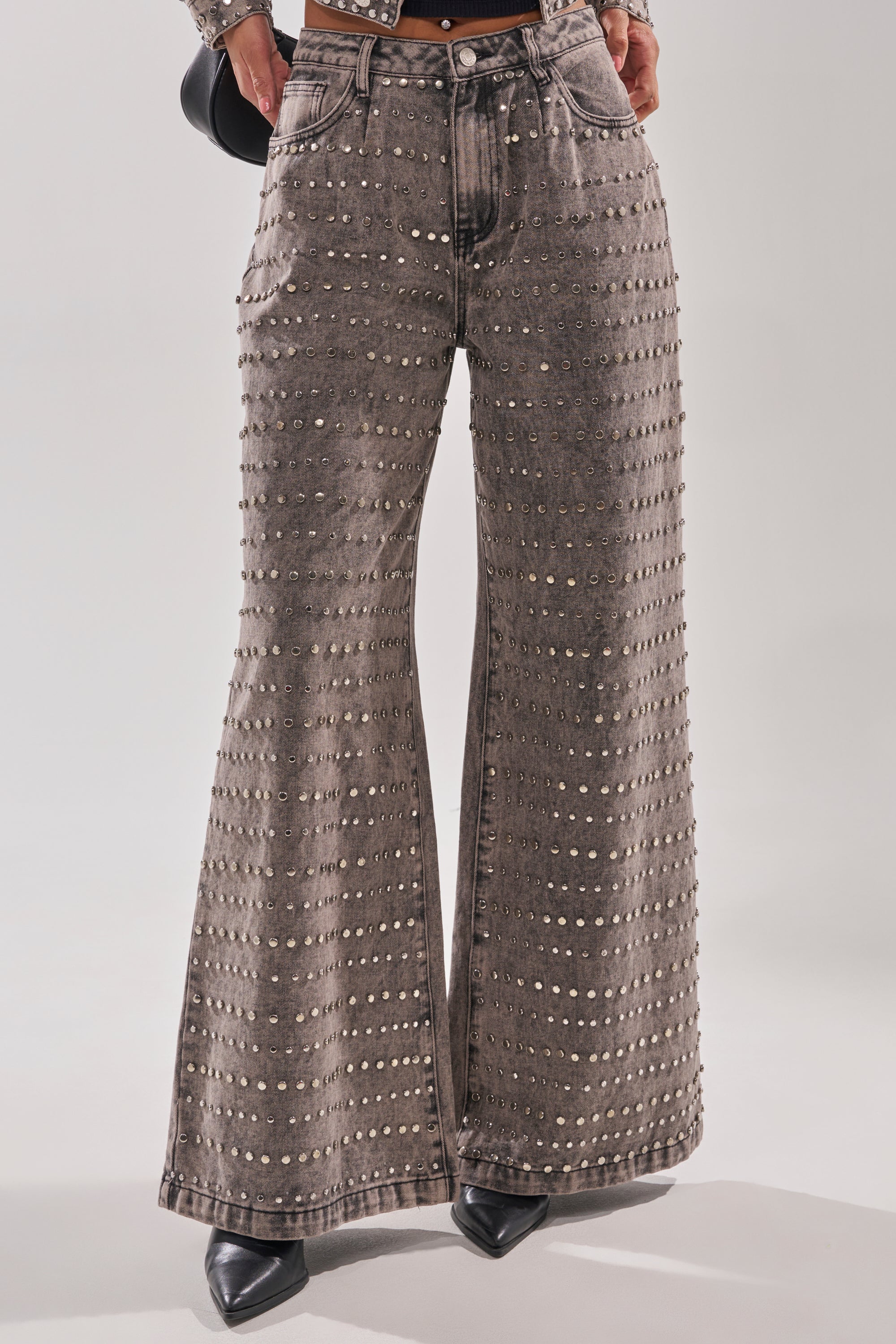 The RIZZO STUDDED WIDE LEG DENIM features a faded gray wash, front pockets, and rows of metallic studs. Styled with pointed black shoes, the photo is cropped to show the high waist jeans and lower body only.
