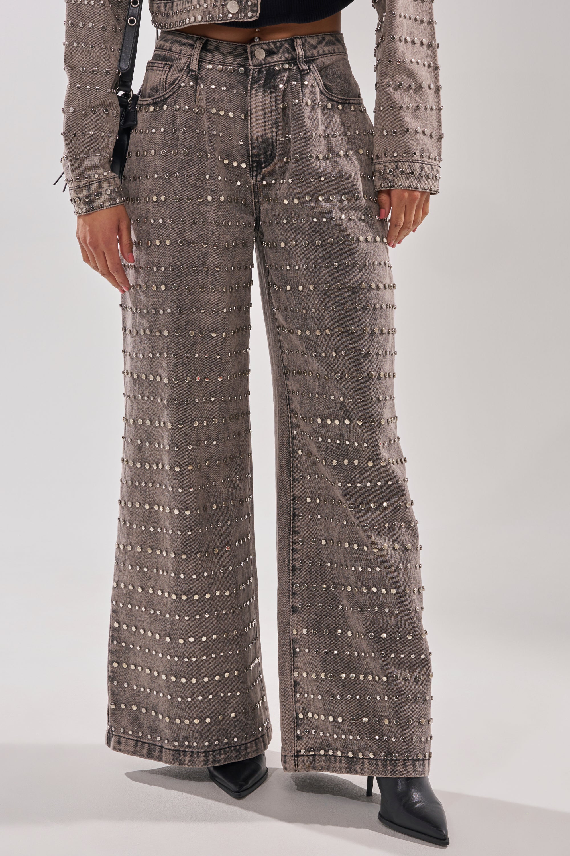 Wearing the RIZZO STUDDED WIDE LEG DENIM with rows of metallic studs, a person pairs them with a matching studded jacket and black pointed-toe boots, standing against a light background.