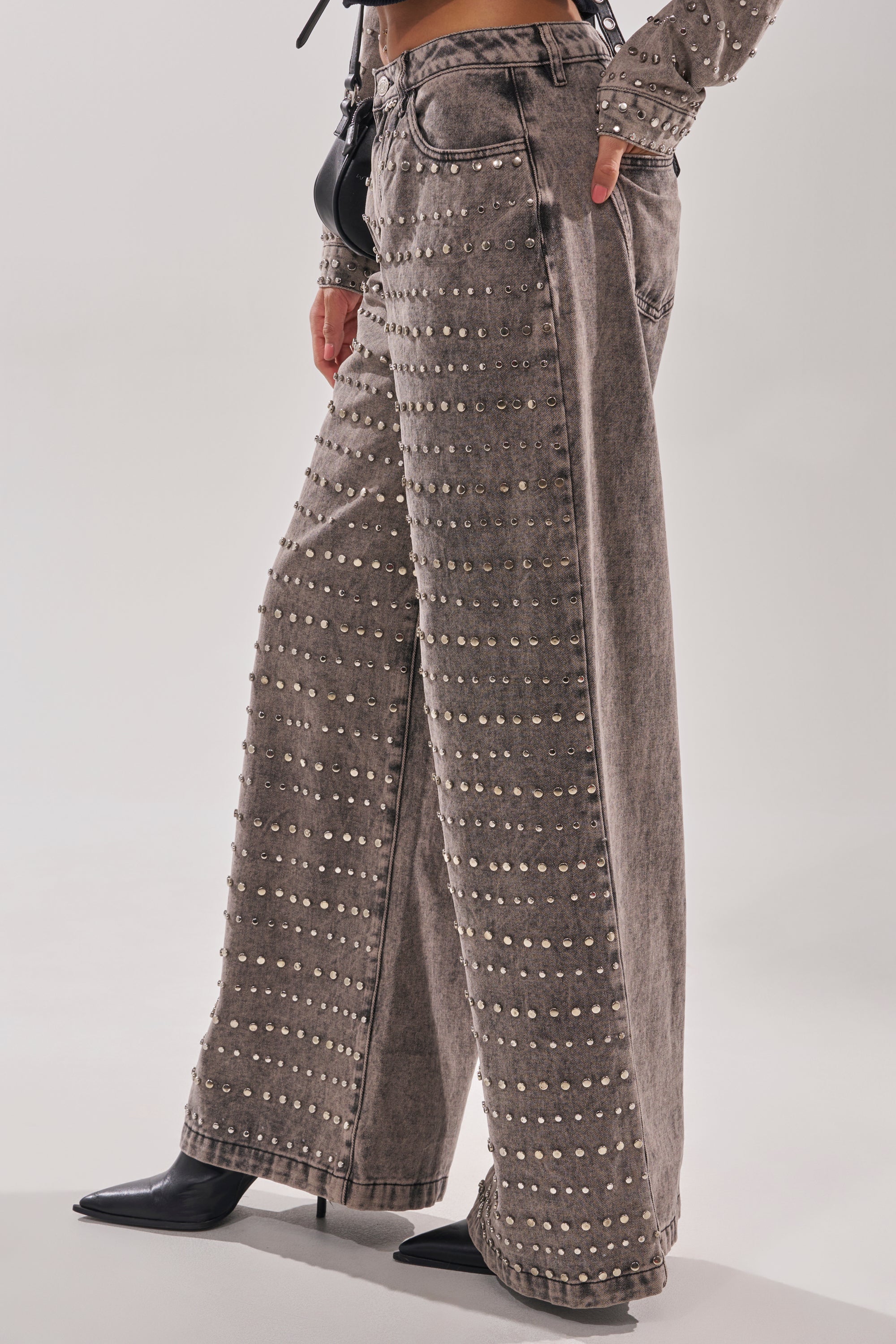 Someone wears the RIZZO STUDDED WIDE LEG DENIM in faded gray with rows of silver studs, paired with black pointed-toe shoes. The upper part of the outfit is partially visible.