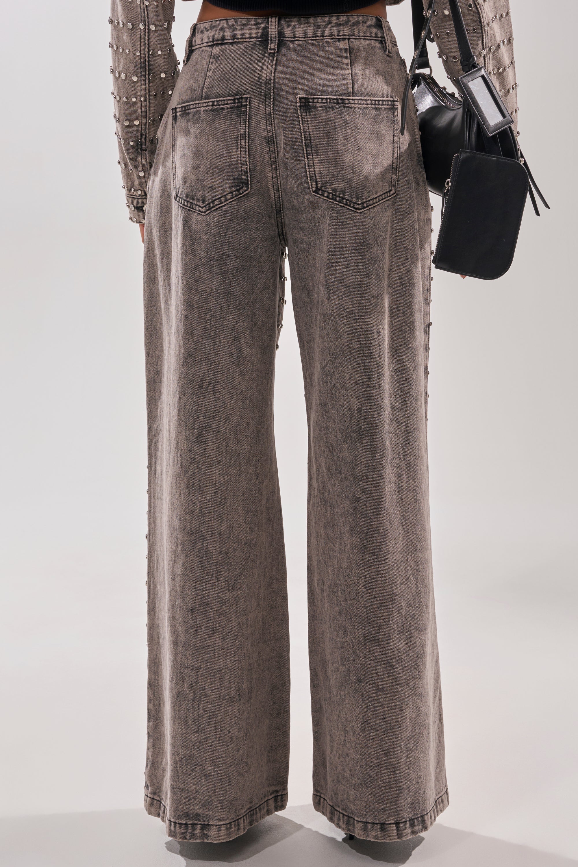 A person shown from the back wears RIZZO STUDDED WIDE LEG DENIM jeans with stud details, a sheer pearl-embellished long-sleeve top, and holds a small black handbag against a plain background.