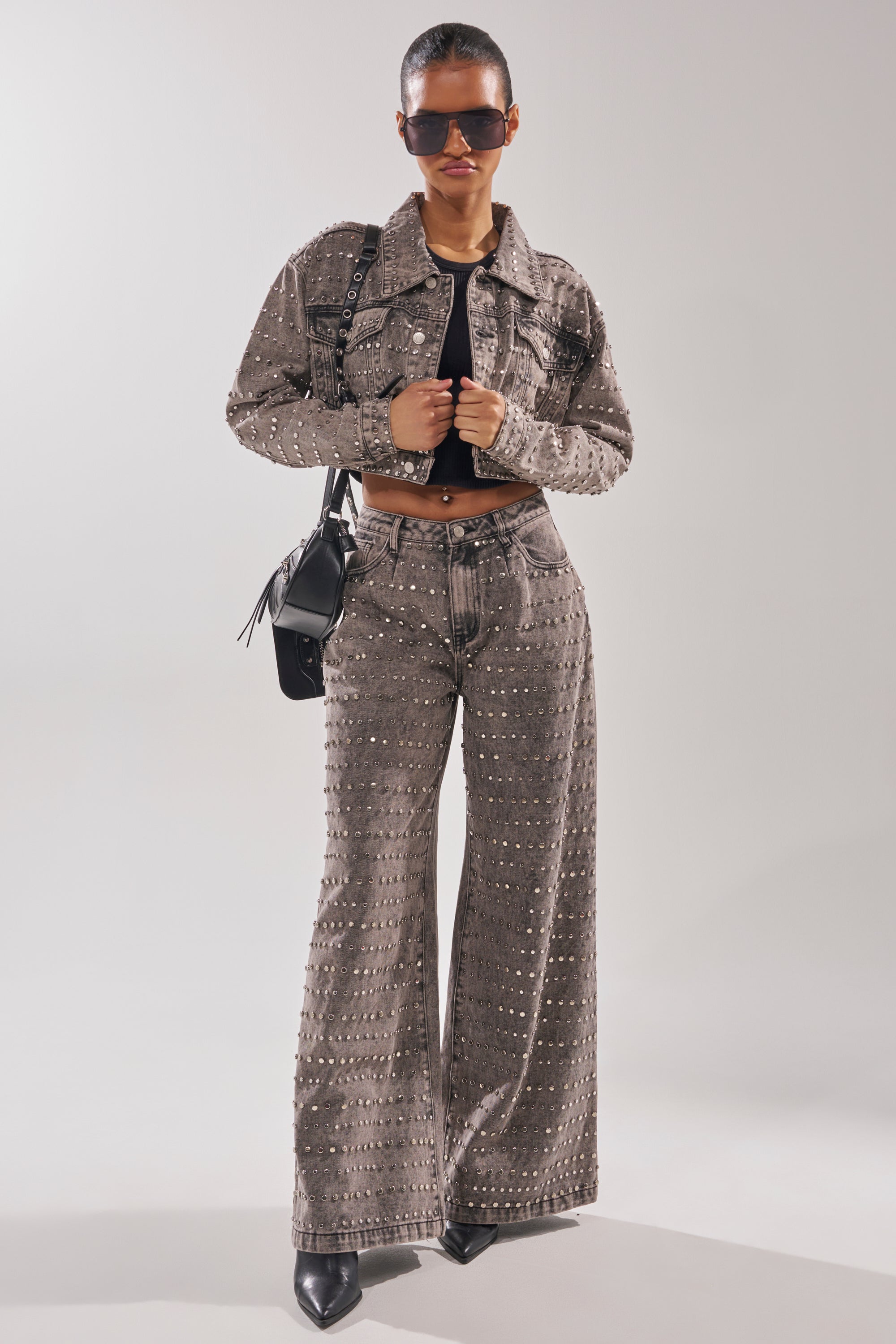 Wearing the RIZZO STUDDED WIDE LEG DENIM—a gray jacket and matching pants—with a black crop top, pointed shoes, sunglasses, and a black crossbody bag, someone stands confidently against a plain light background.