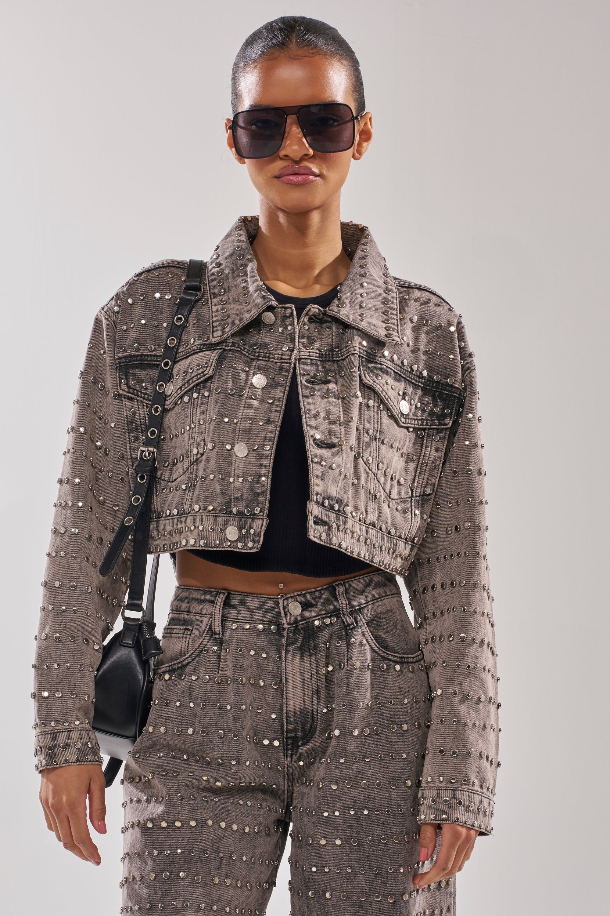 A woman channels rocker-chic style, wearing large sunglasses, the RIZZO STUDDED DENIM JACKET with matching high-waisted pants, a black crop top, and a black shoulder bag against a plain background.