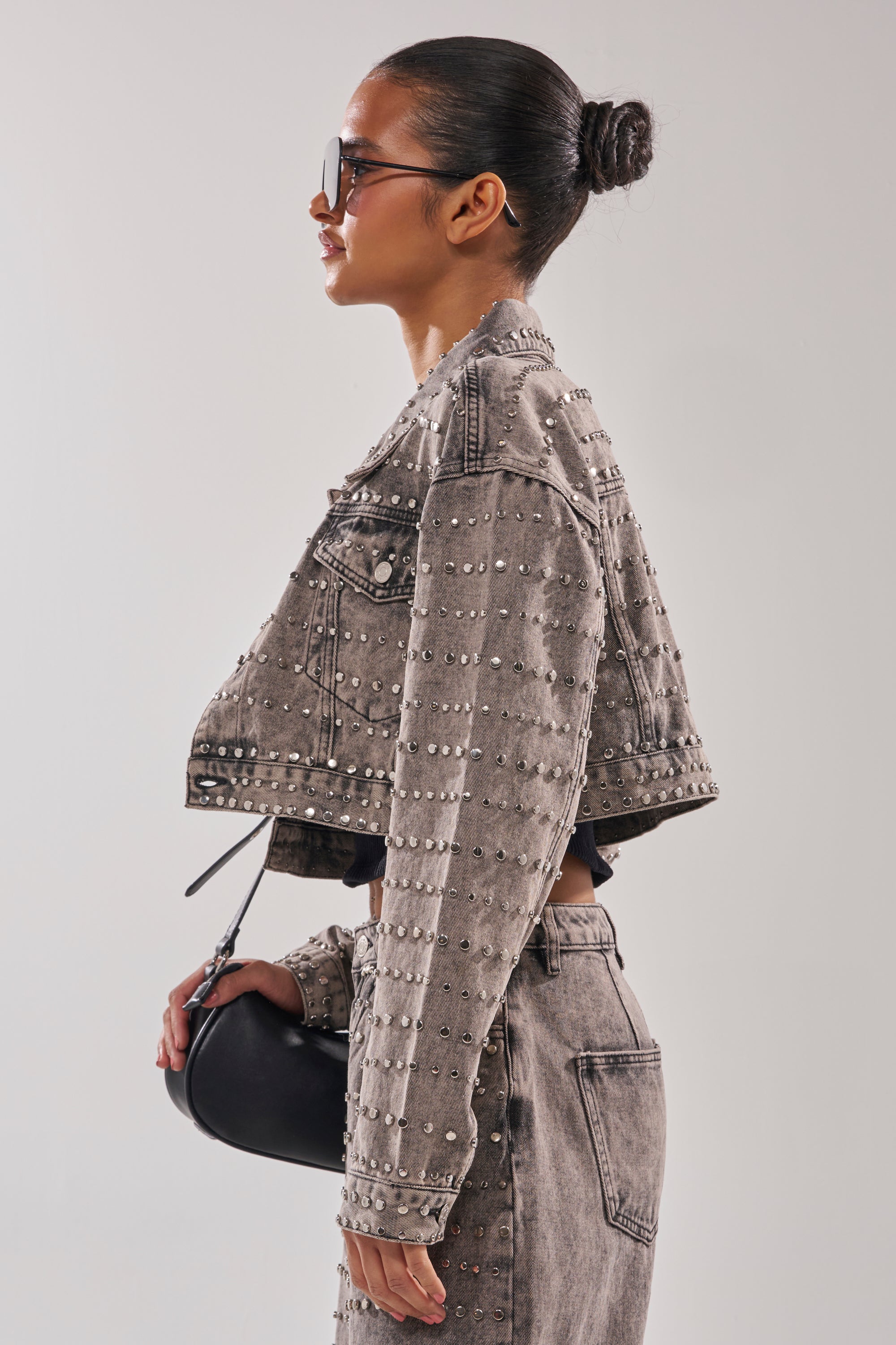 A woman in profile wears sunglasses, the RIZZO STUDDED DENIM JACKET, and matching high-waisted jeans. She carries a small black handbag, her hair in a neat bun, set against a plain light background.