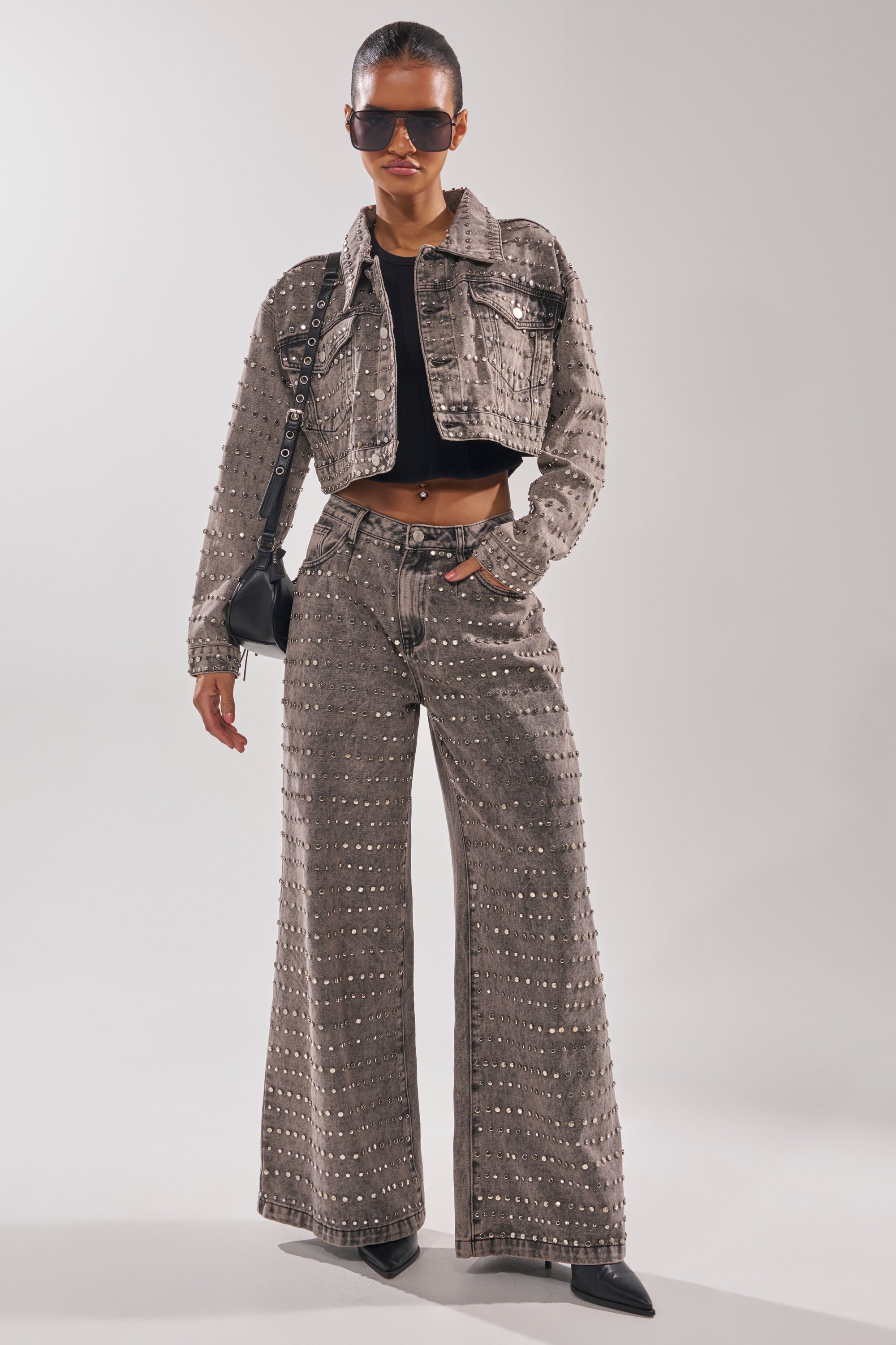 A woman stands confidently against a plain light background in the RIZZO STUDDED DENIM JACKET, matching gray wide-leg pants, a black crop top, pointed black shoes, large sunglasses, and a black shoulder bag.