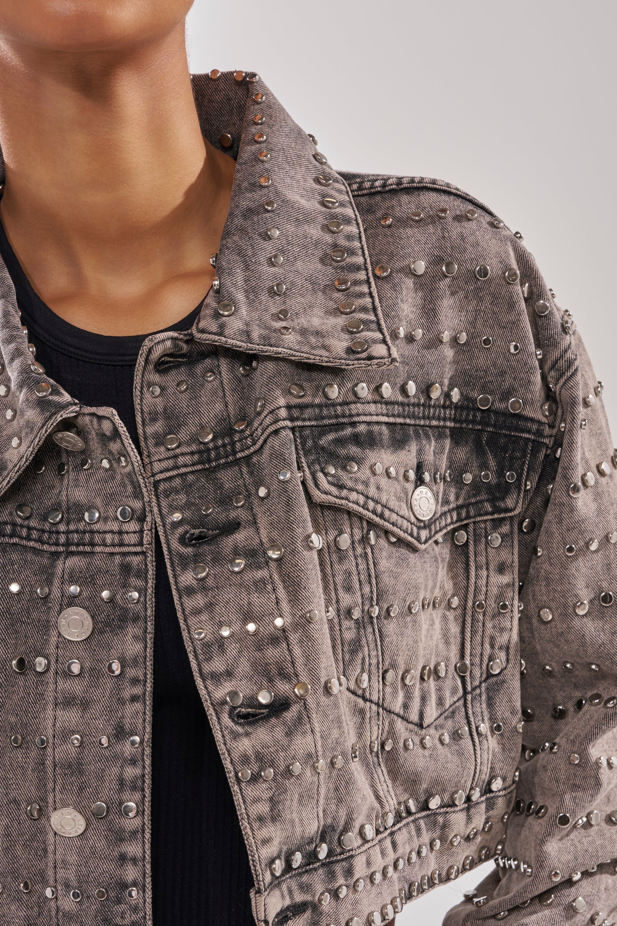 A close-up of a person in the RIZZO STUDDED DENIM JACKET, featuring gray acid-wash denim, studded details, and a chest pocket, styled over a black ribbed top for a rocker-chic look.