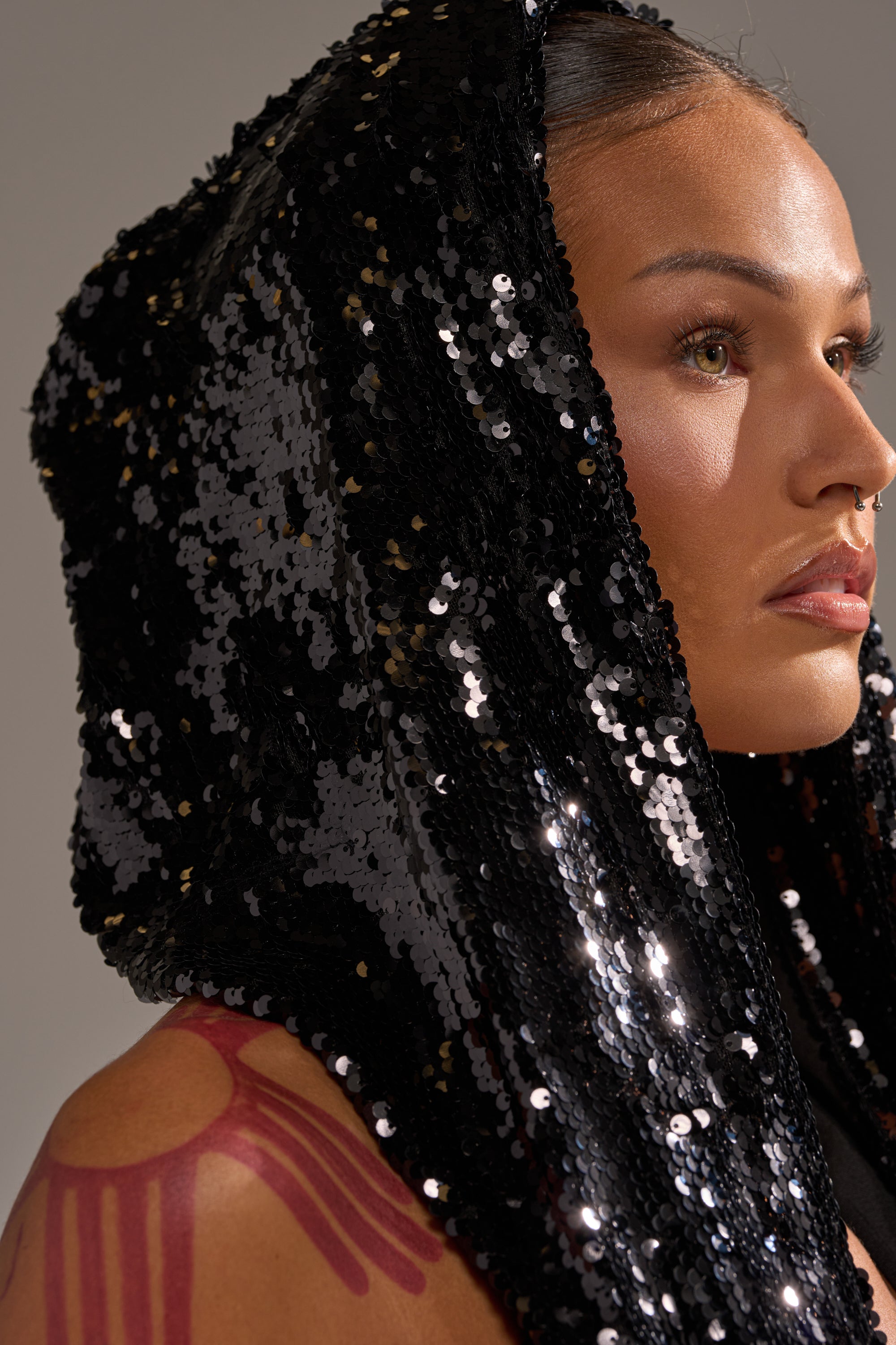 Wearing the LOVE ME LIKE THAT SEQUIN MINI DRESS with a black sequin hood, a person with smooth skin, subtle makeup, and a red sun tattoo on their shoulder poses sideways as lighting highlights the sequins and their facial features.