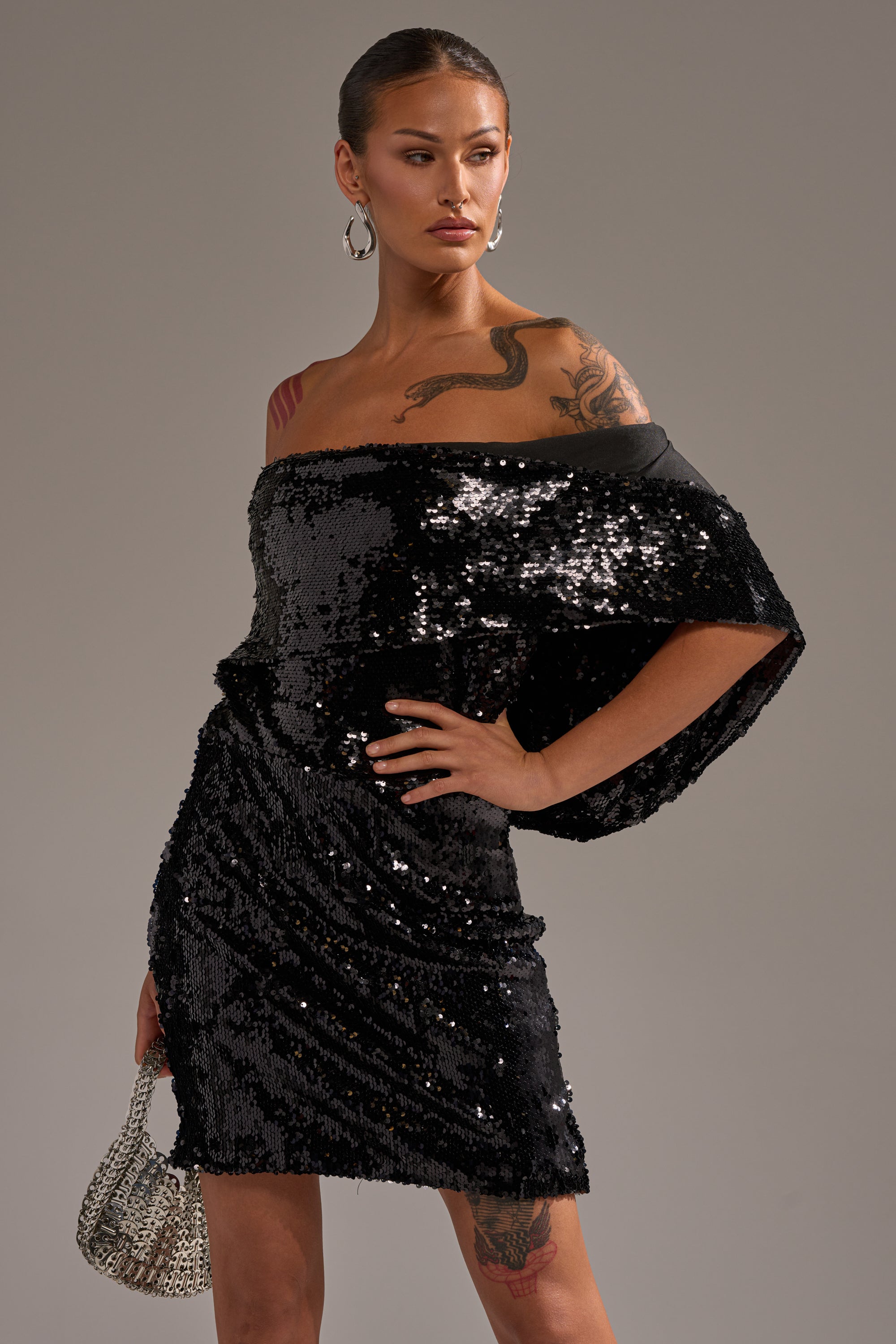 Wearing the LOVE ME LIKE THAT SEQUIN MINI DRESS with a cowl neckline, this person poses confidently with a silver handbag, tattoos on their arm and shoulder, hair pulled back, and large hoop earrings.