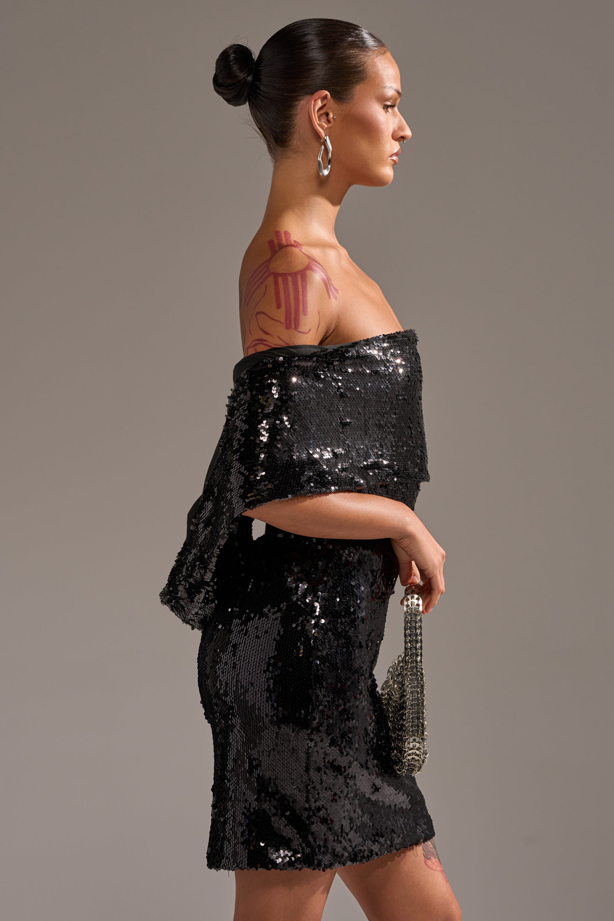 A woman with a bun hairstyle and a large pink shoulder tattoo wears the LOVE ME LIKE THAT SEQUIN MINI DRESS, holding a small purse and facing left against a plain gray background.