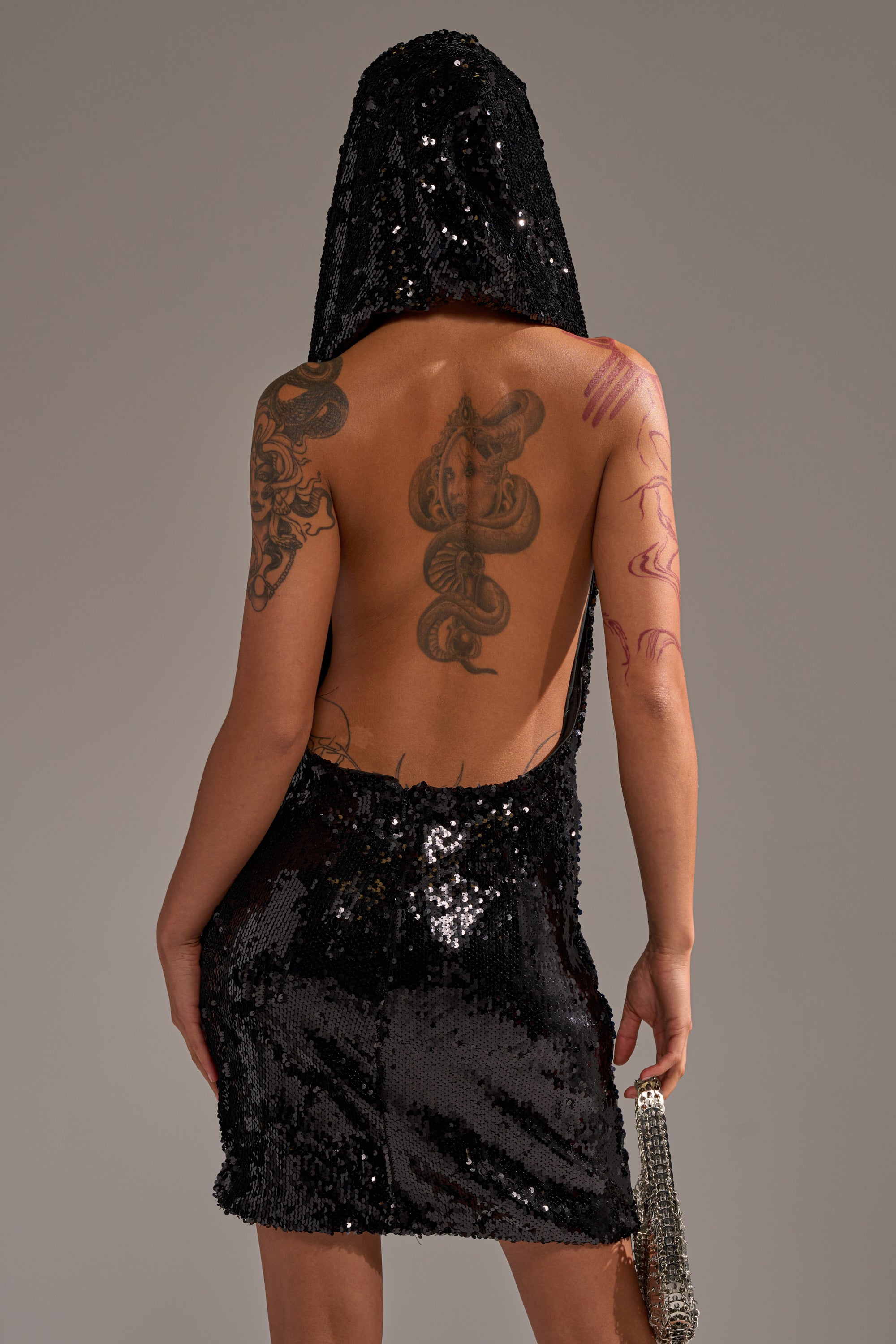 Wearing the LOVE ME LIKE THAT SEQUIN MINI DRESS—a black, backless mini with sequins, cowl neckline, and hood—a person shows tattoos on their arm and back as they face away from the camera, against a gray background with a shiny mini handbag.