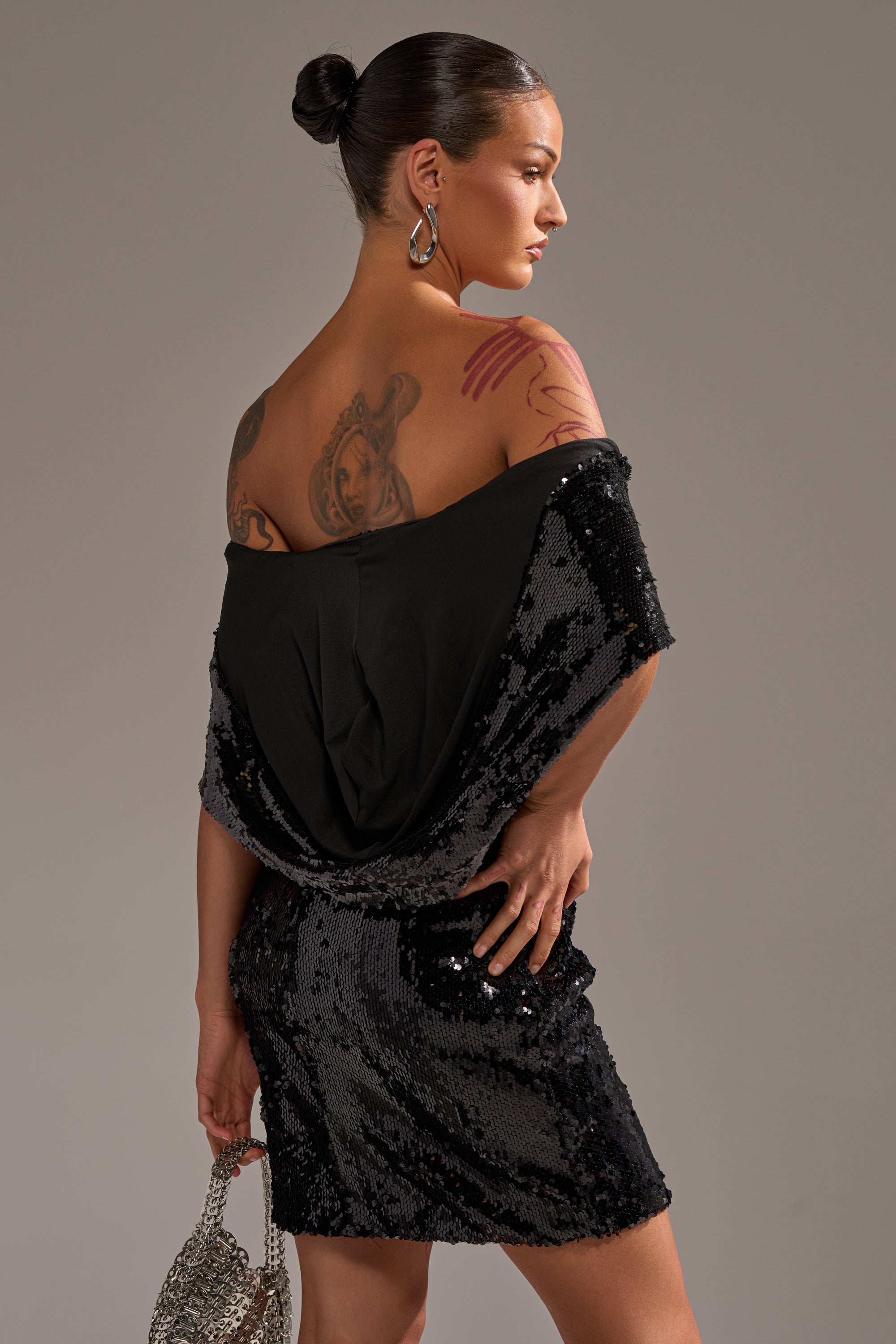 A woman with visible tattoos poses in the LOVE ME LIKE THAT SEQUIN MINI DRESS, featuring an off-shoulder design. Her hair is styled in a sleek bun as she holds a small silver purse and gazes to the side against a neutral background.