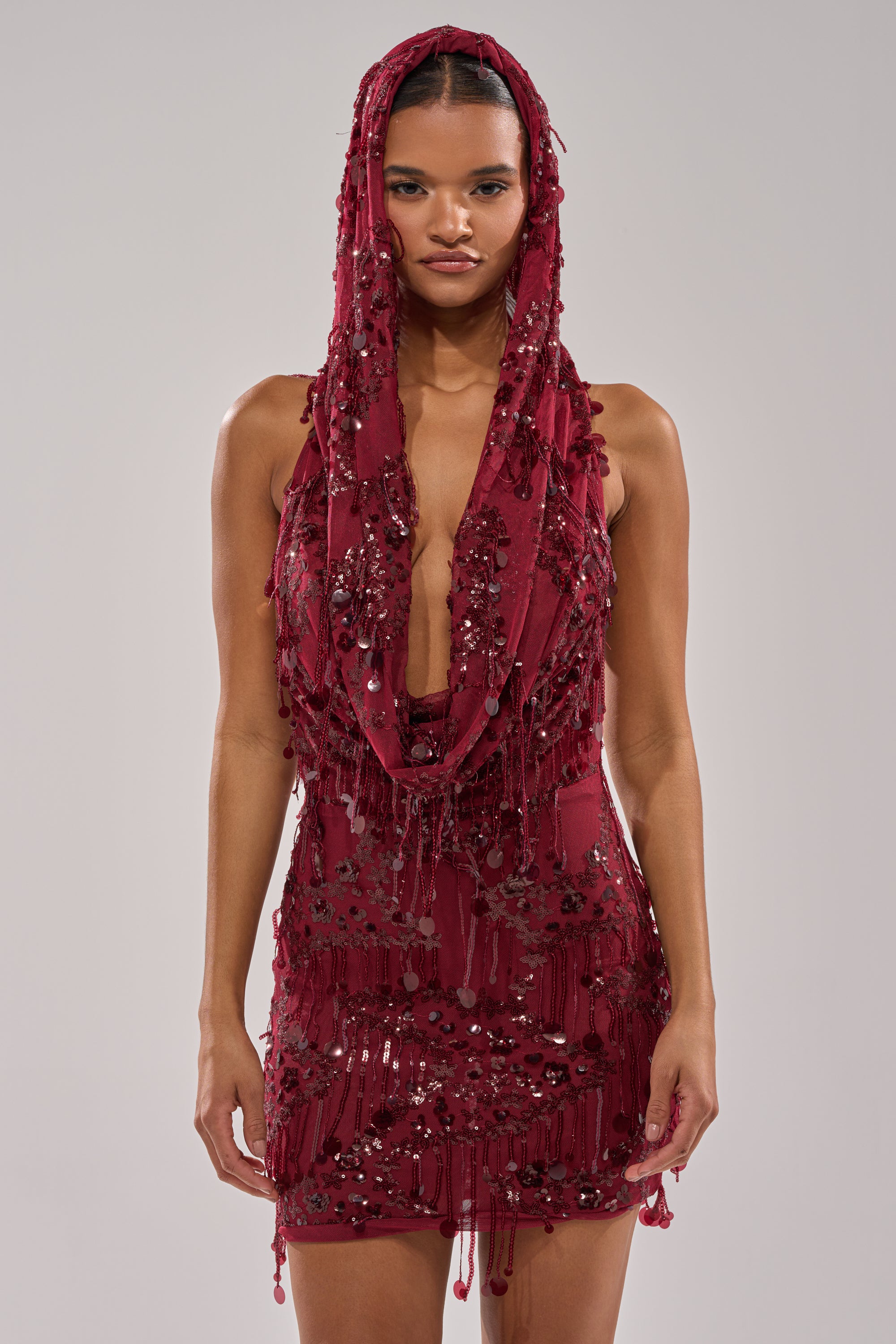 A woman wears the MOOD FOR THE NIGHT SEQUIN MINI DRESS, a sleeveless wine red mini with a dramatic cowl hood, beadwork, and fringe detail, standing against a plain light gray background.