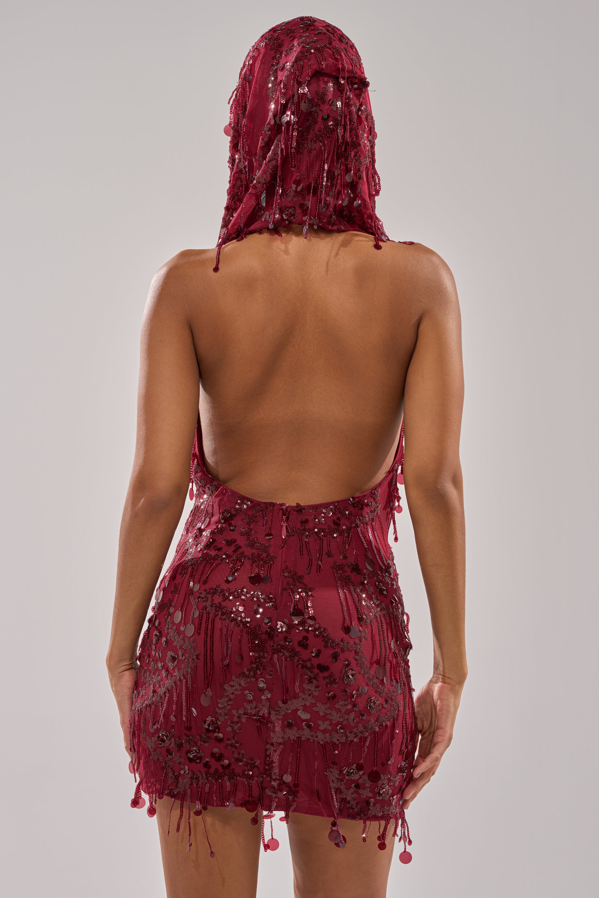 A person stands with their back to the camera, wearing the MOOD FOR THE NIGHT SEQUIN MINI DRESS in wine red, featuring a matching hood, shimmering sequins, beads, an off-the-shoulder design, and open back against a plain light background.