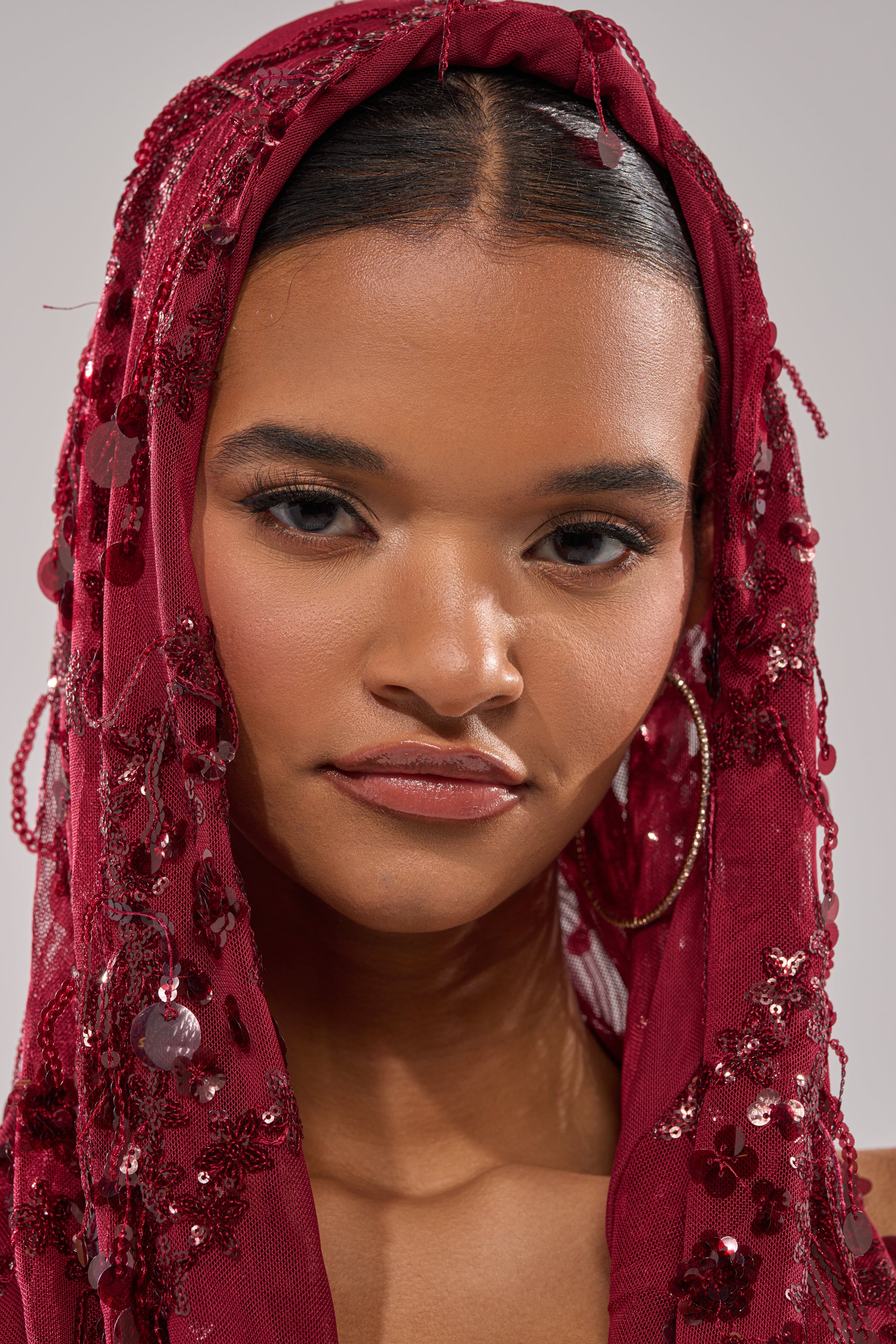 A woman with smooth medium-brown skin and a calm, confident expression wears the MOOD FOR THE NIGHT SEQUIN MINI DRESS with an off-the-shoulder cut, a red beaded and sequined headscarf, hoop earrings, and subtle makeup.