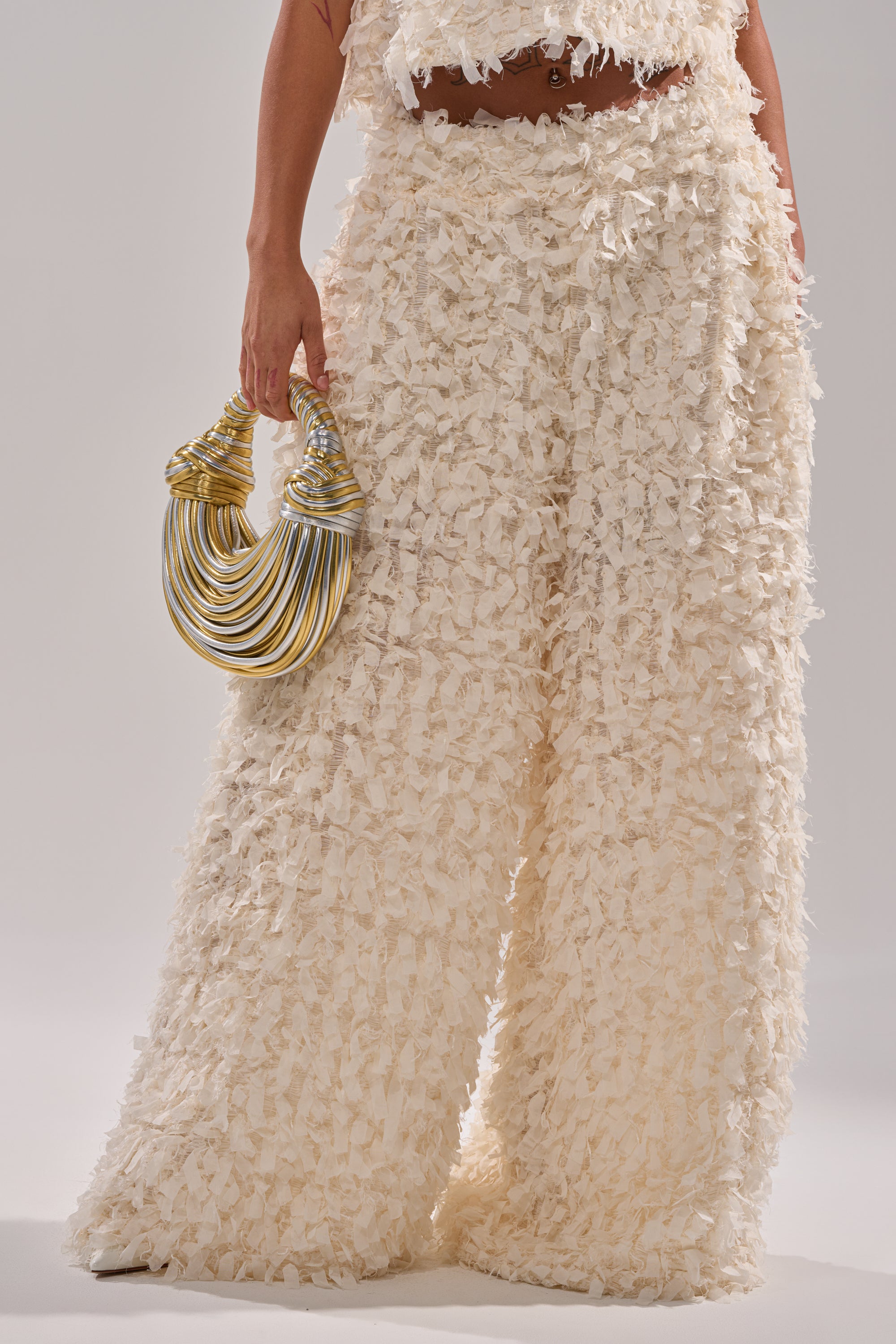 Wearing the MOXIE WIDE LEG TROUSER in cream textured fabric with a matching top, a person holds a gold and silver sculptural handbag. Only the lower torso, part of one arm, and the bag appear against a plain background. 1