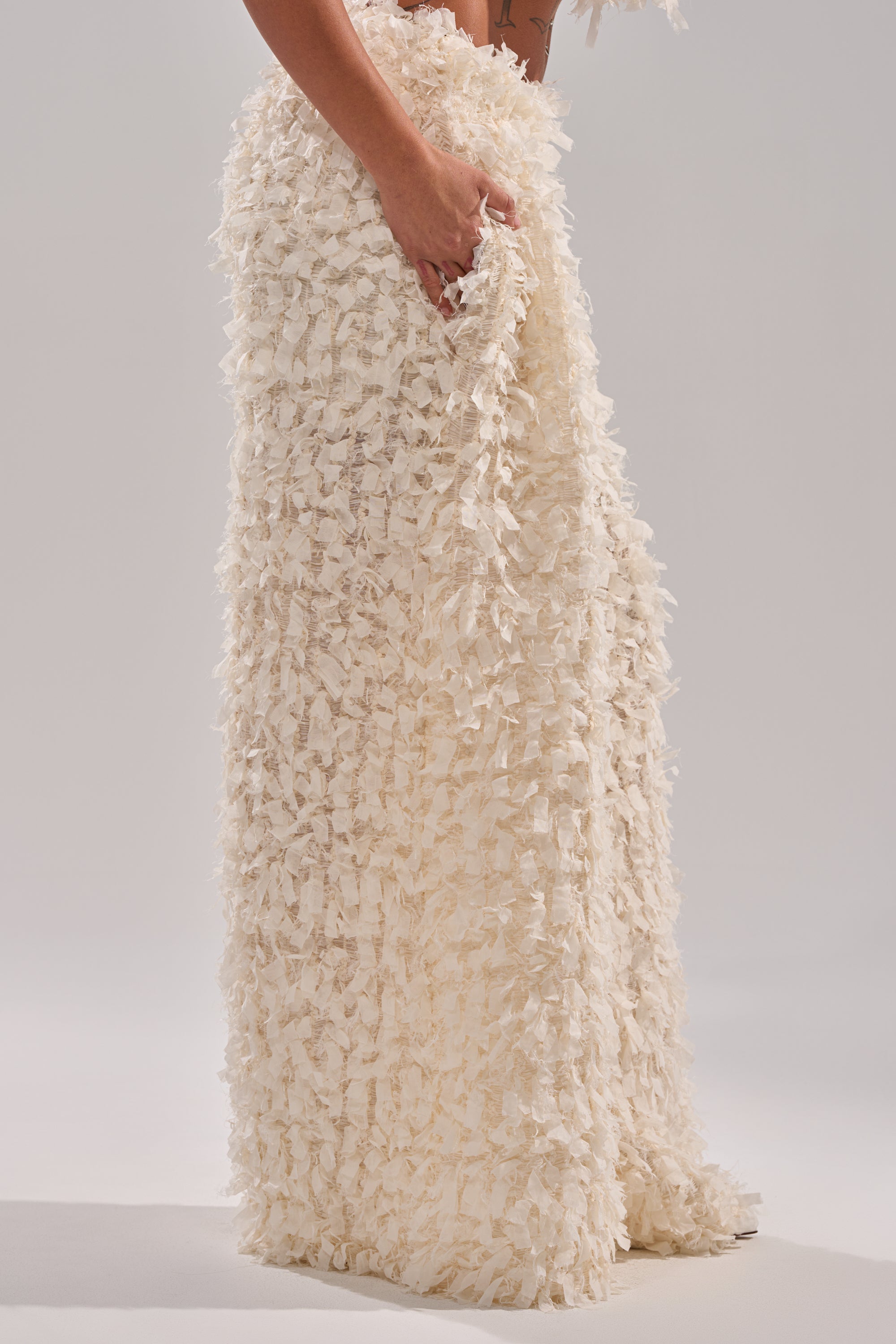 A person stands against a plain light background, wearing the MOXIE WIDE LEG TROUSER—an ivory, wide-leg pant featuring layered, fringed mesh fabric—resting one hand on the trouser. 2