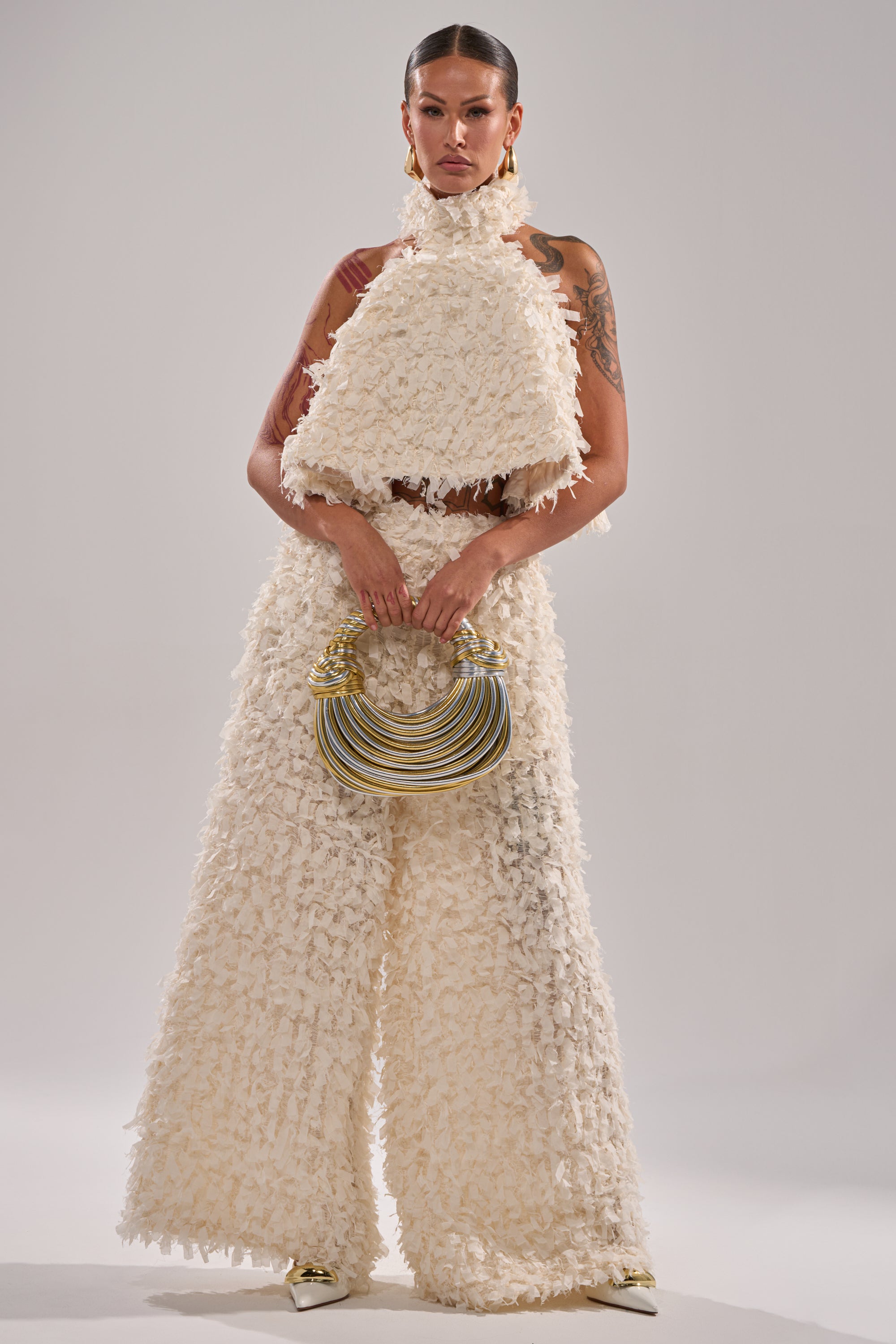 A woman wears the MOXIE WIDE LEG TROUSER with a cream sleeveless top, pointed shoes, and holds a small handbag with gold and silver hoops. Her hair is slicked back, and she shows off her tattoos. 5
