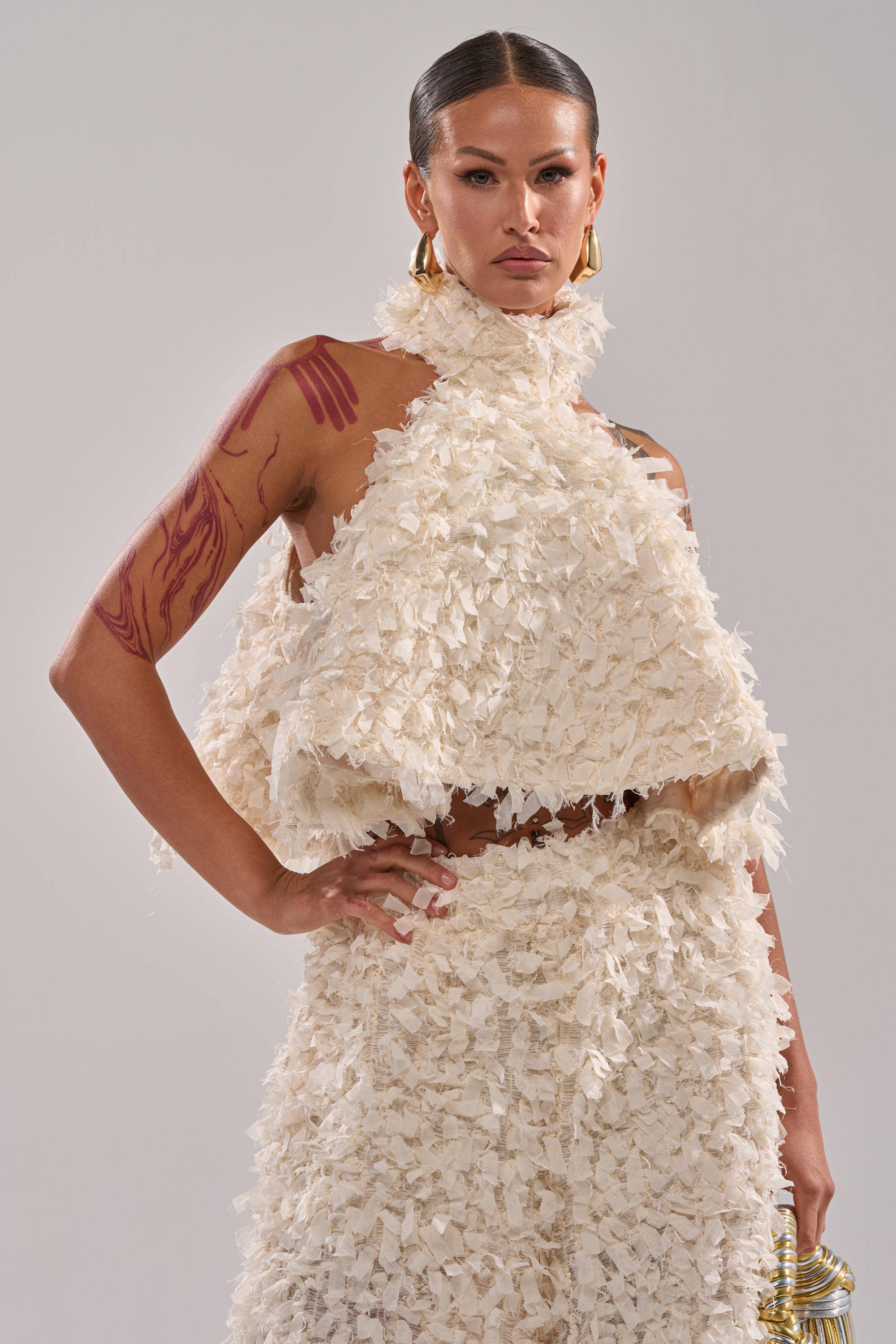 A model with slicked-back hair wears the MOXIE HALTER NECK BLOUSE in off-white, featuring ruffled and ruched details. Red tattoos on her left arm and gold hoop earrings complete her confident pose, one hand resting on her hip. 0