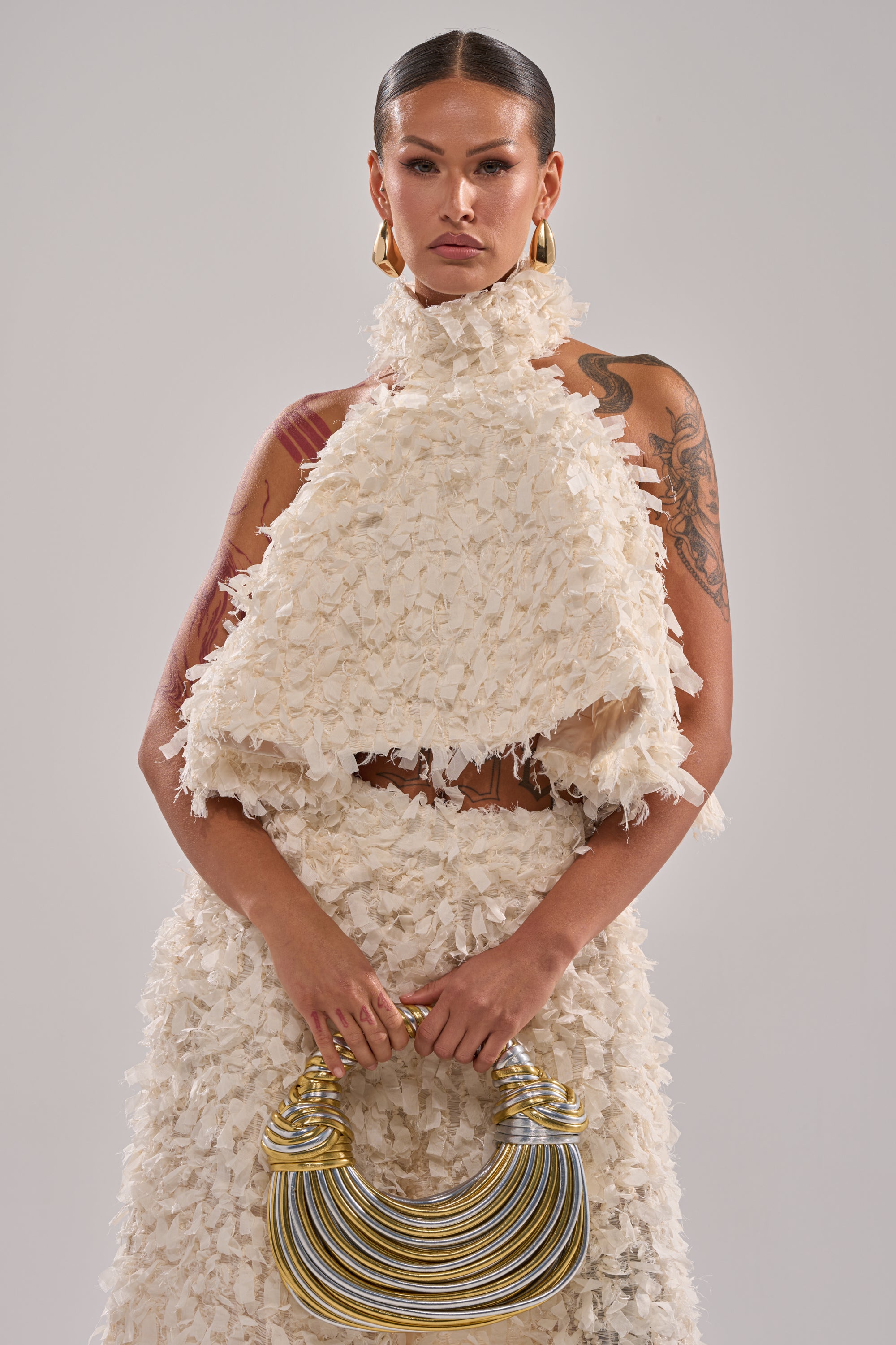 A woman with slicked-back hair and tattoos wears an off-white MOXIE HALTER NECK BLOUSE and skirt, gold hoop earrings, and holds a sculptural gold-and-silver handbag while standing against a plain background. 1