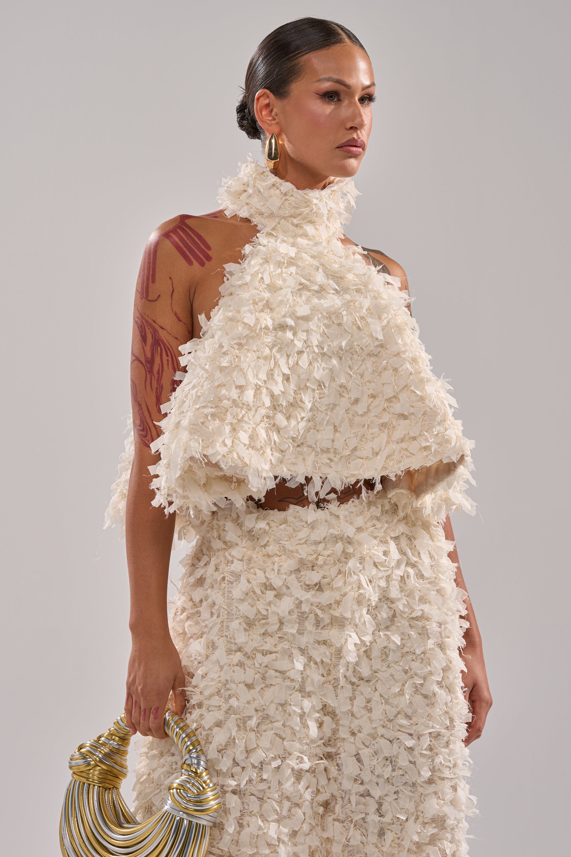 A woman with slicked-back hair wears the white MOXIE HALTER NECK BLOUSE and matching skirt. She holds a gold sculptural bag, showing arm tattoos, against a plain light gray background. 2