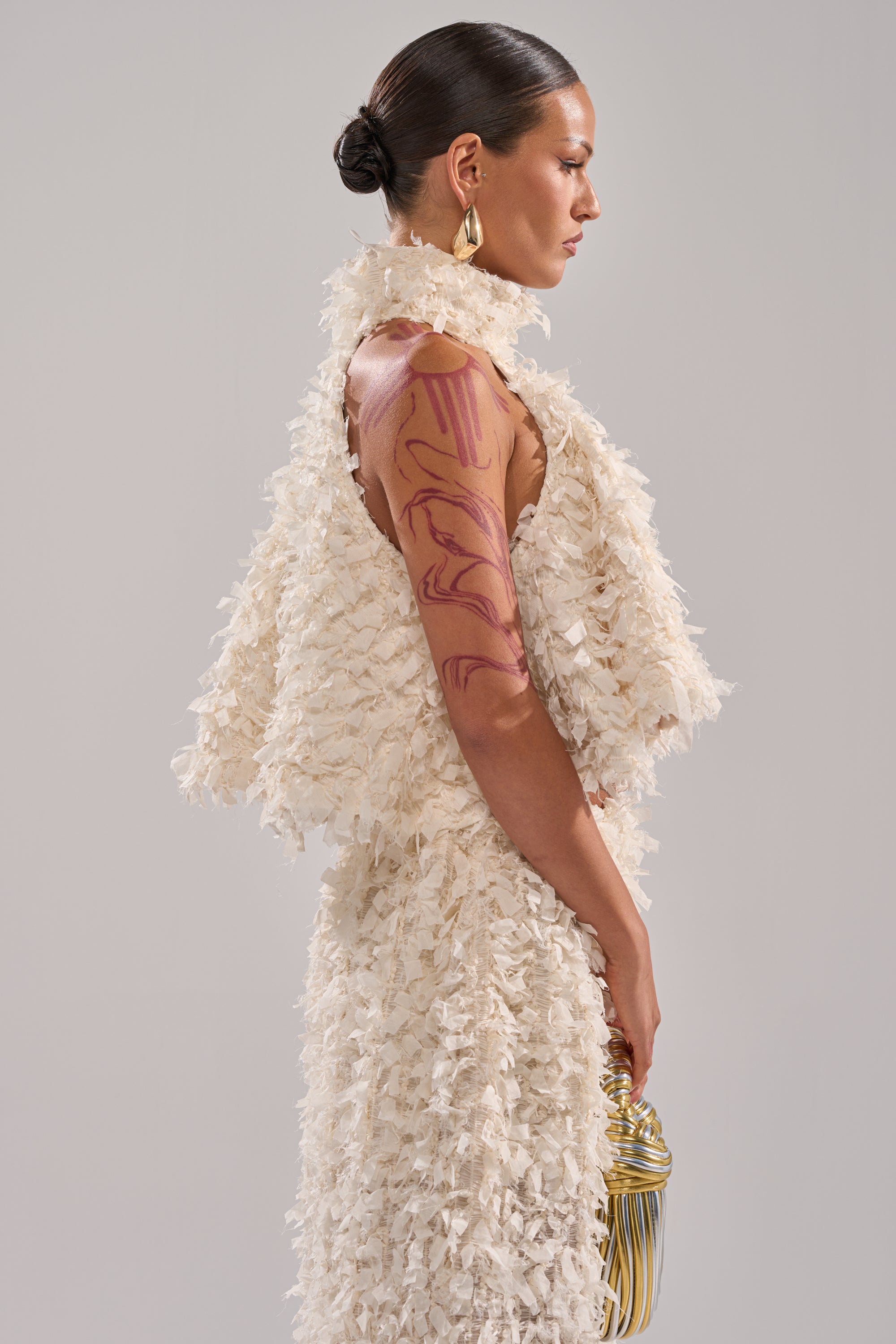A woman in profile wears the off-white MOXIE HALTER NECK BLOUSE with a matching vest, styled with a sleek bun, large gold earrings, visible red tattoos on her upper arm, and holding a gold metallic clutch. 3