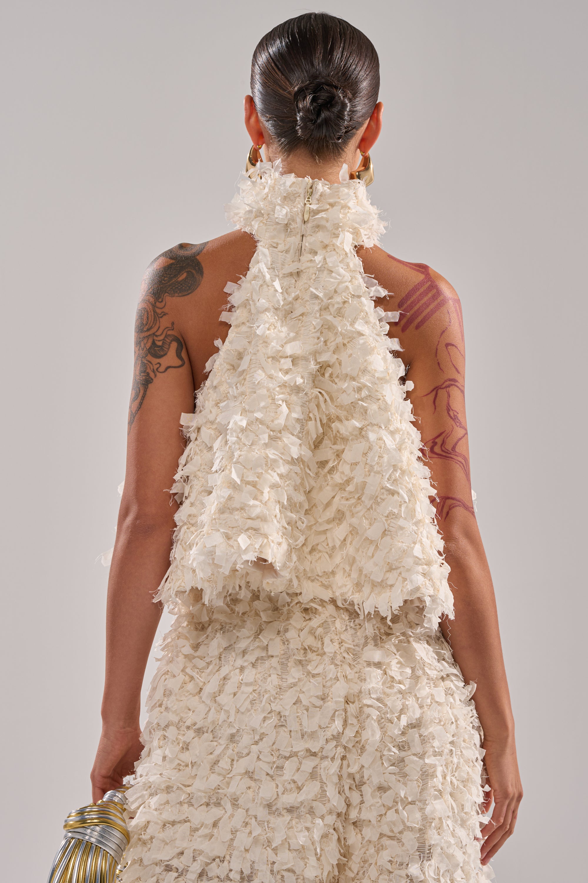 A person with slicked-back hair and visible tattoos is seen from behind against a plain background, wearing the MOXIE HALTER NECK BLOUSE—a textured white halter neck style with a high collar—and holding a metallic handbag. 4