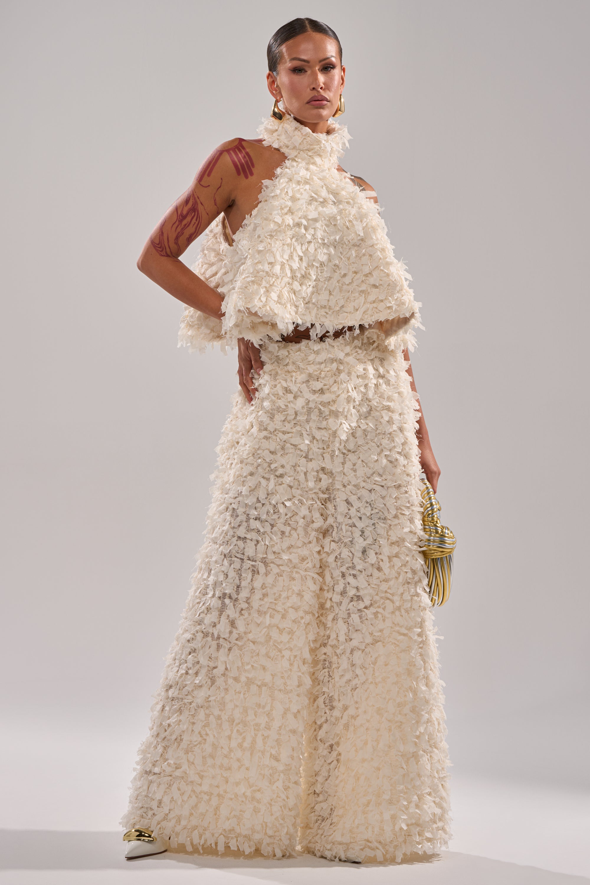 Wearing the MOXIE HALTER NECK BLOUSE and wide-leg pants, a person poses confidently, holding a small gold handbag with bold earrings, slicked-back hair, and arm tattoos on display. 5